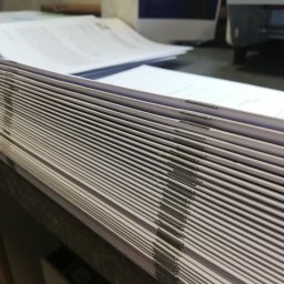 Powielarnia MADAS - Stos zszytych katalogów lub broszur z widocznymi zszywkami, leżący na ciemnej powierzchni, z rozmazanym tłem zawierającym dokumenty i fragment maszyny.