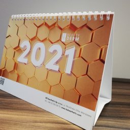 Powielarnia MADAS - Stojący kalendarz biurkowy spiralowany, z grafiką złotych heksagonów i logo Beauty Derm Aesthetic, rok 2021, umieszczony na drewnianym blacie.