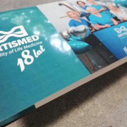 Powielarnia MADAS - Błyszczący katalog firmy Artismed z okazji 18-lecia, z zespołem medycznym w turkusowych strojach i balonami, leżący na szarej powierzchni.