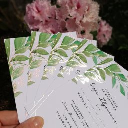 Powielarnia MADAS - Stos zaproszeń na sesję zdjęciową z motywem zielonych liści i złotych akcentów, trzymany w dłoni na tle kwitnącego krzewu.