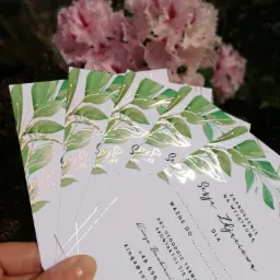 Stos zaproszeń na sesję zdjęciową z motywem zielonych liści i złotych akcentów, trzymany w dłoni na tle kwitnącego krzewu.