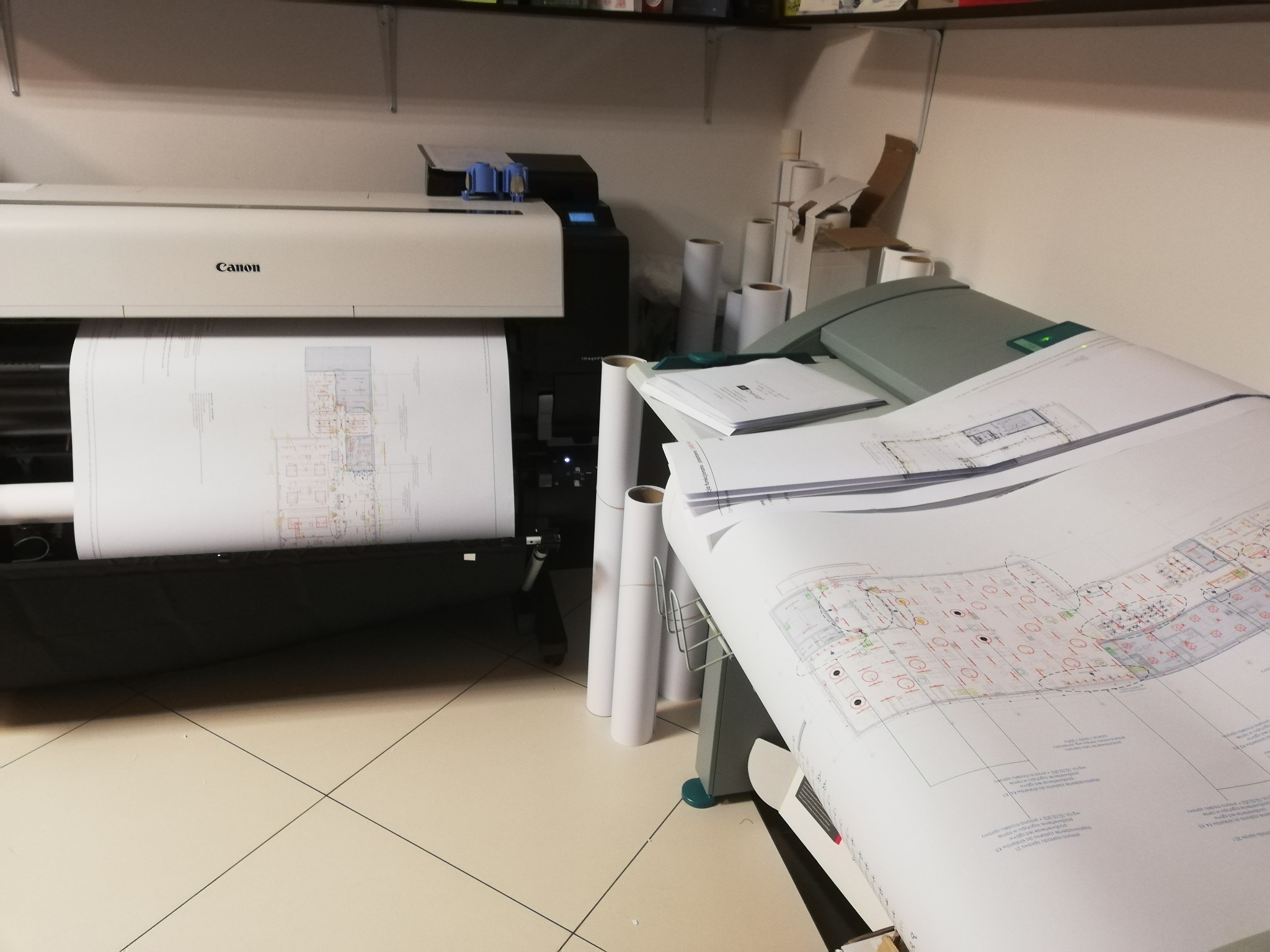 Szerokoformatowa drukarka Canon w trakcie drukowania planu architektonicznego; obok rolki papieru i wydrukowane plany.