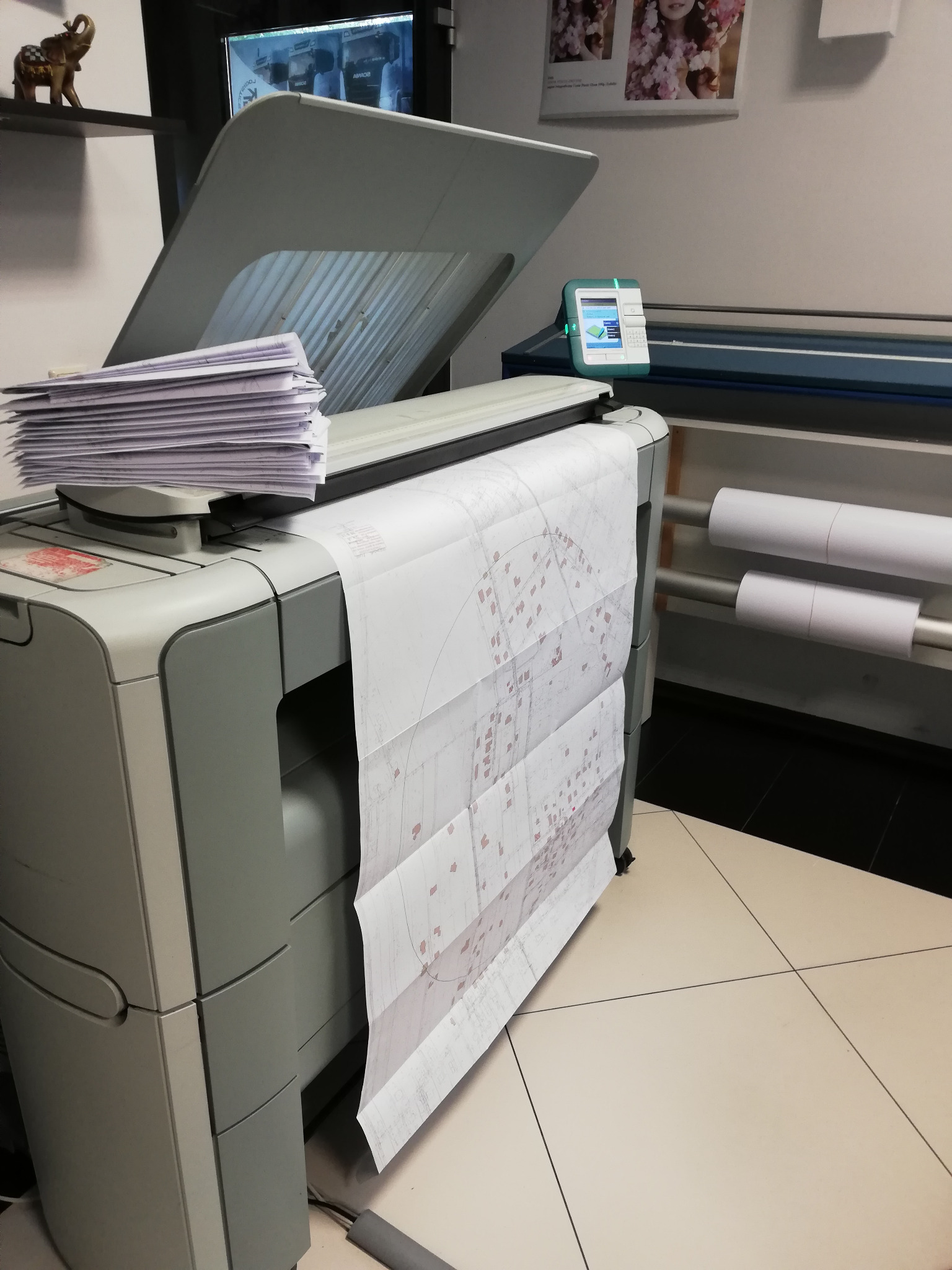 Skaner wielkoformatowy z otwartą klapą, skanujący duży plan architektoniczny, sterta dokumentów na górze, rolki papieru w tle.