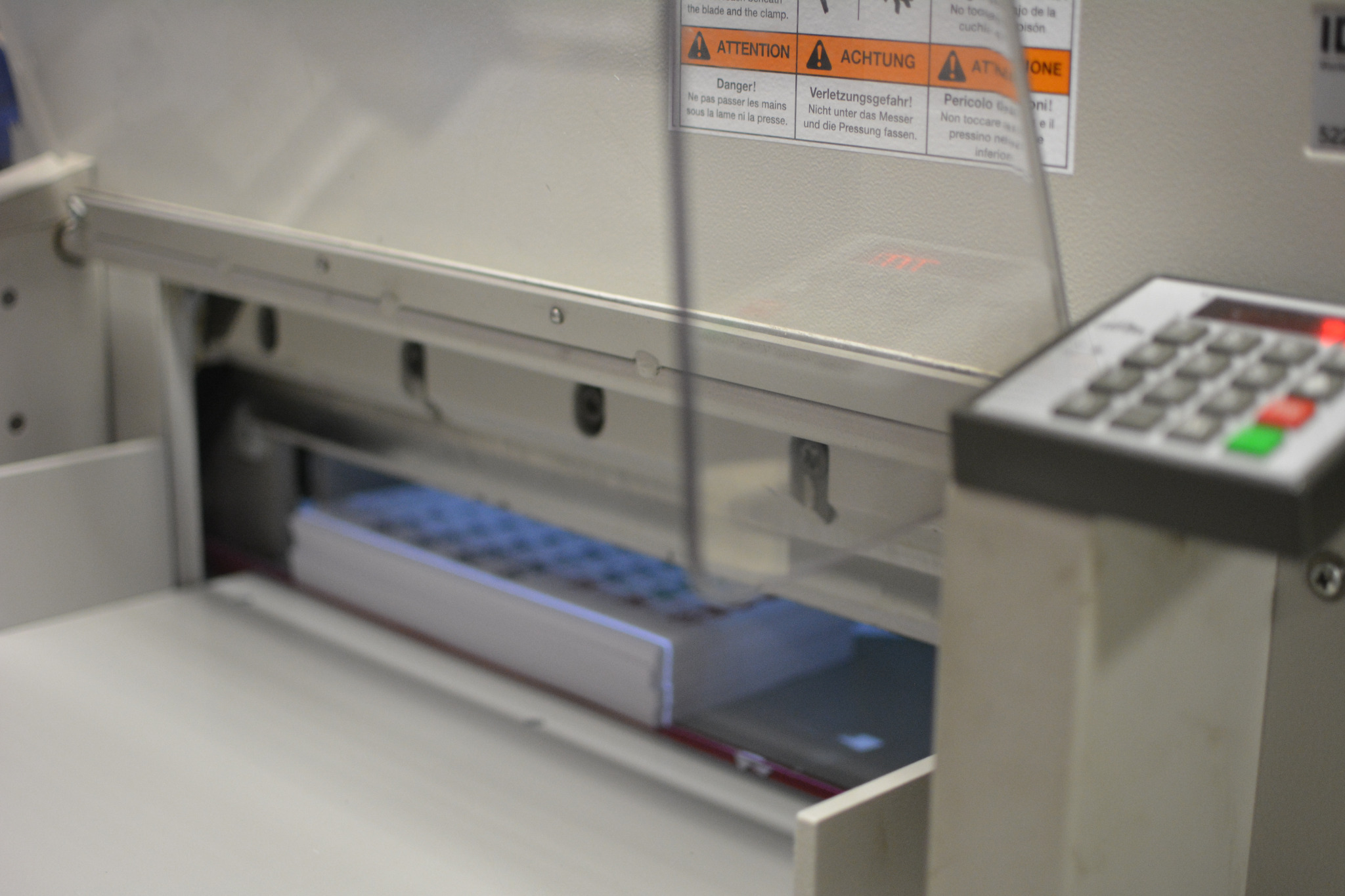 Gilotyna introligatorska z stosem papieru wewnątrz, widoczne ostrze i panel sterowania z przyciskami, naklejki ostrzegawcze na obudowie.
