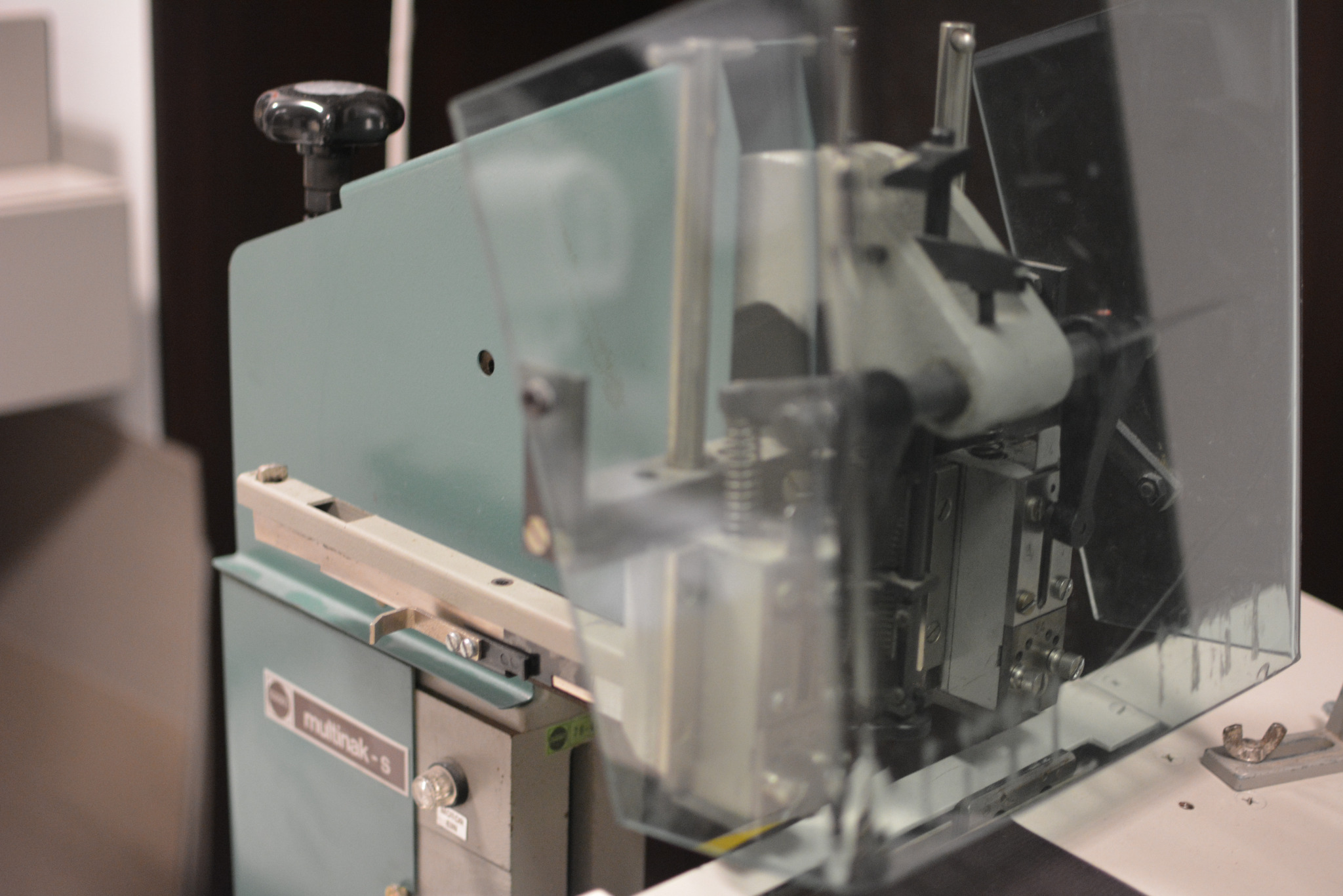 Maszyna introligatorska Multinak-S z przezroczystą osłoną, widoczne mechanizmy i elementy regulacyjne, oświetlenie studyjne.