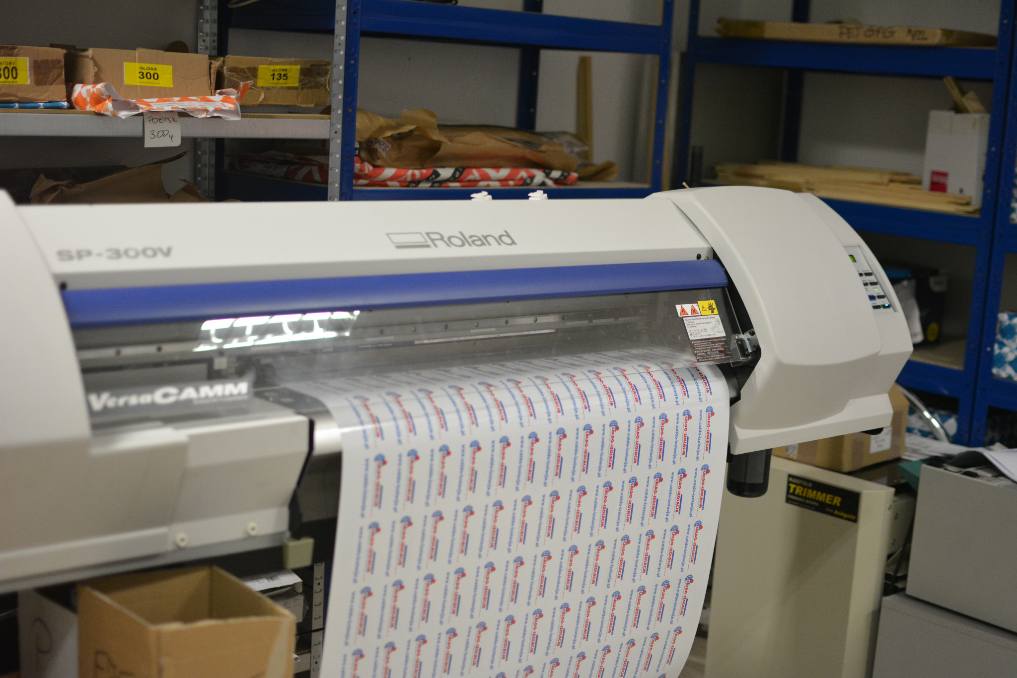 Drukarka wielkoformatowa Roland VersaCAMM SP-300V w trakcie drukowania arkusza z powtarzalnym wzorem logo i adresem strony internetowej firmy Caldo-Izolacja, widoczne półki magazynowe w tle.