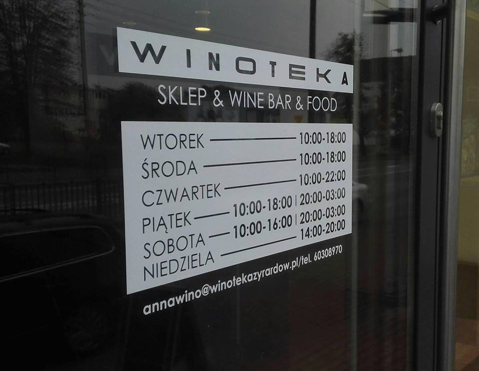 Godziny otwarcia sklepu z winami i delikatesami 'Winoteka' naklejone na szklanych drzwiach wejściowych, widoczne odbicie otoczenia w szybie.