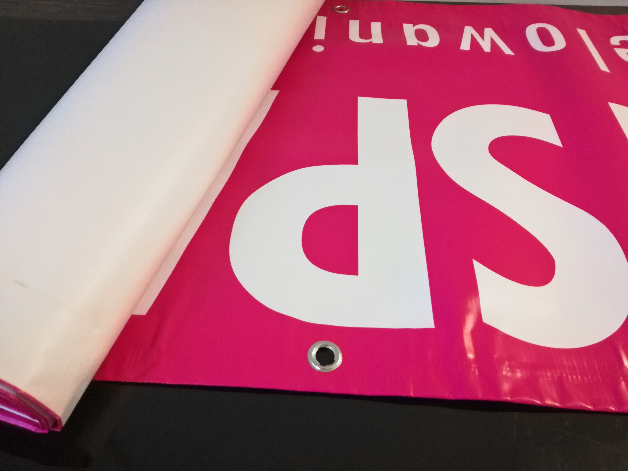 Fragment różowego baneru z białym napisem 'SP' i oczkiem montażowym, częściowo zakryty przez białą rolkę materiału.