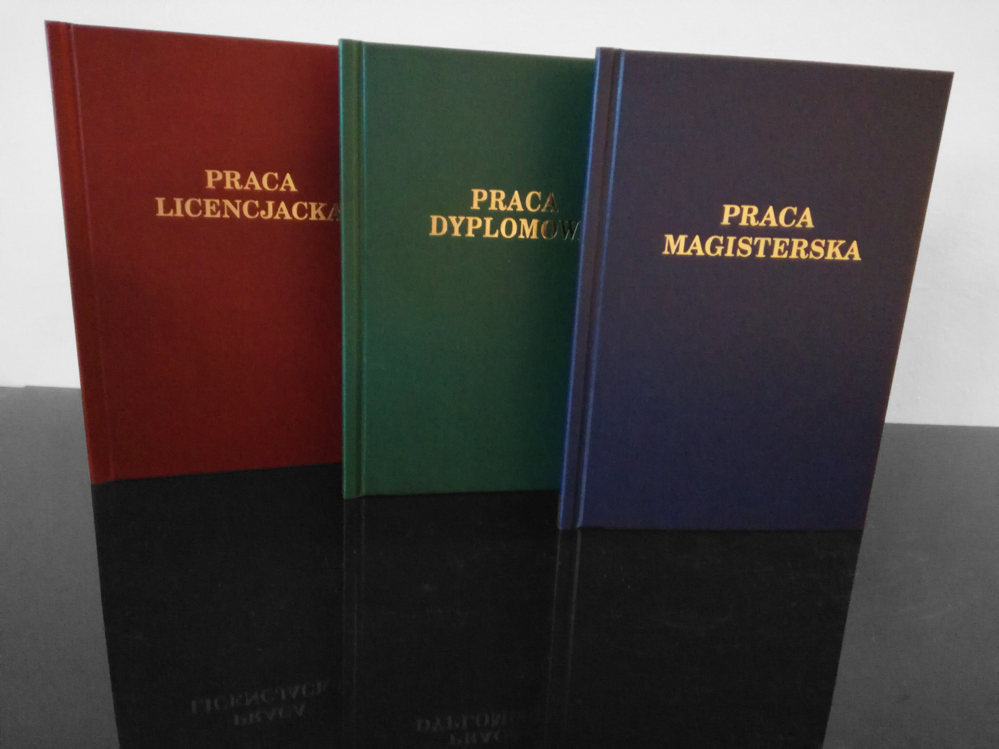 Oprawione prace dyplomowe: licencjacka w bordowej oprawie, magisterska w granatowej i dyplomowa w zielonej, z tłoczonym złotym napisem na okładkach, ustawione rzędem na błyszczącej powierzchni.