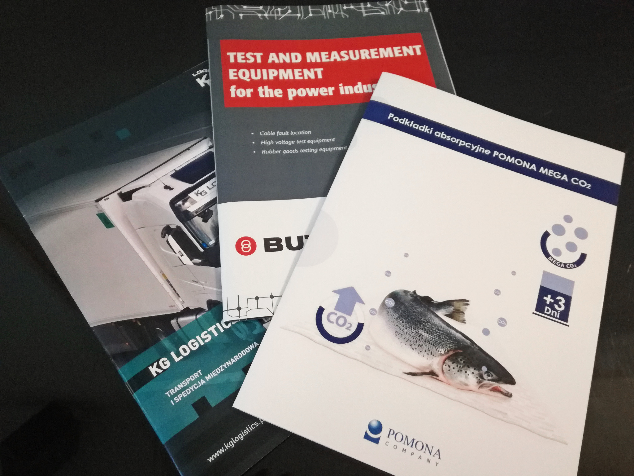 Stos ulotek i broszur firmowych, w tym ulotka firmy transportowej KG Logistics, broszura z wyposażeniem pomiarowym i ulotka o podkładkach absorpcyjnych Pomona Mega CO2 z grafiką ryby.