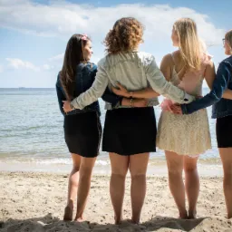 Cztery kobiety stojące tyłem na plaży, objęte ramionami, w tle morze i niebo.