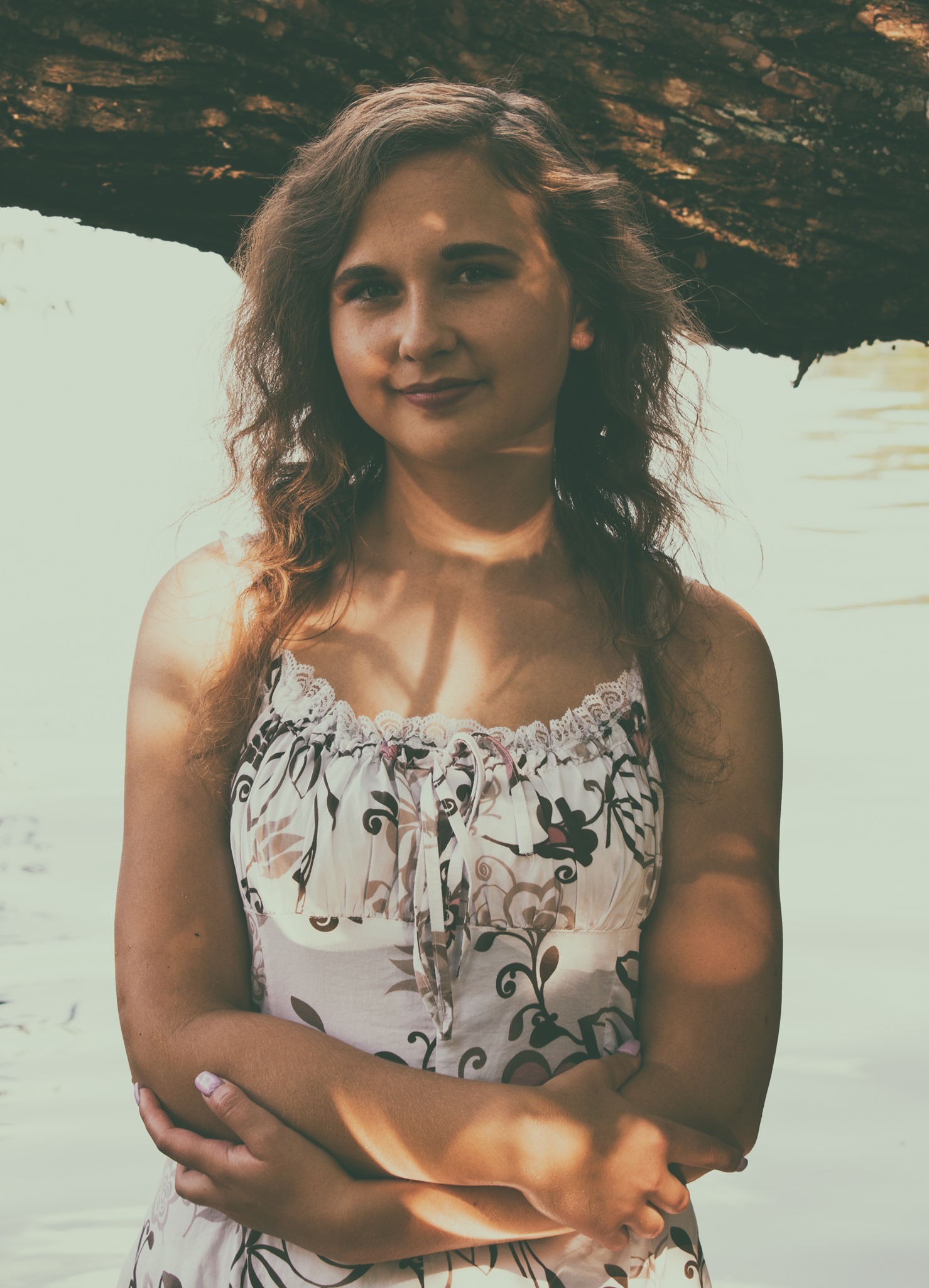 Portret młodej kobiety w letniej sukience z kwiatowym wzorem, stojącej pod gałęzią drzewa w naturalnym świetle.