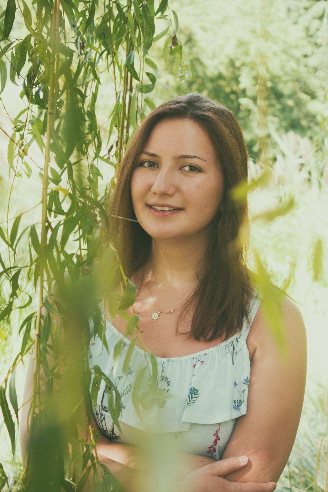 Uśmiechnięta kobieta w kwiecistej bluzce, pozująca na tle zieleni drzew z delikatnym naszyjnikiem.