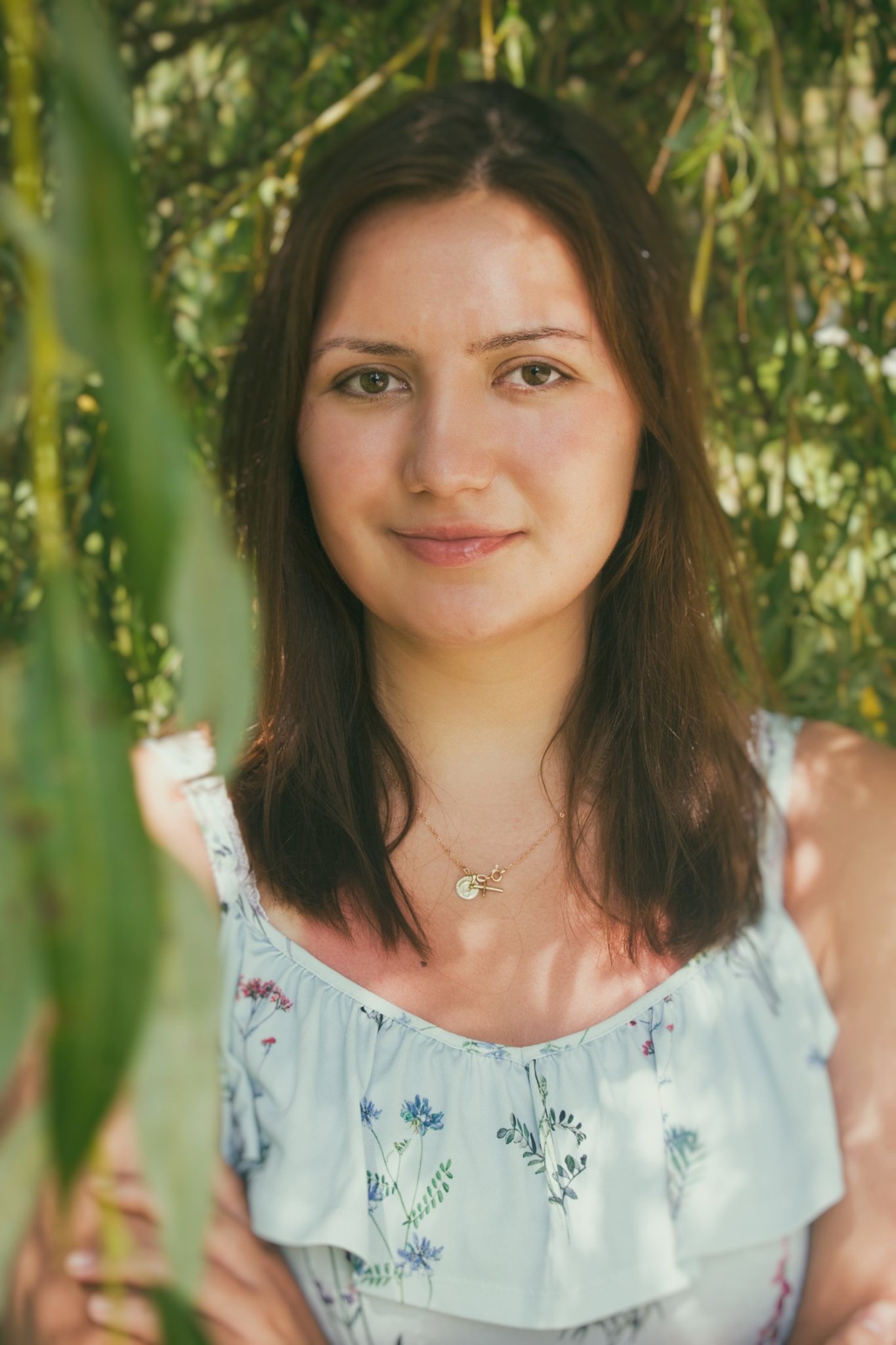 Portret młodej kobiety z delikatnym uśmiechem, ubrana w bluzkę w kwiaty, pozuje na tle zieleni z wiszącymi liśćmi, złoty naszyjnik z zawieszką