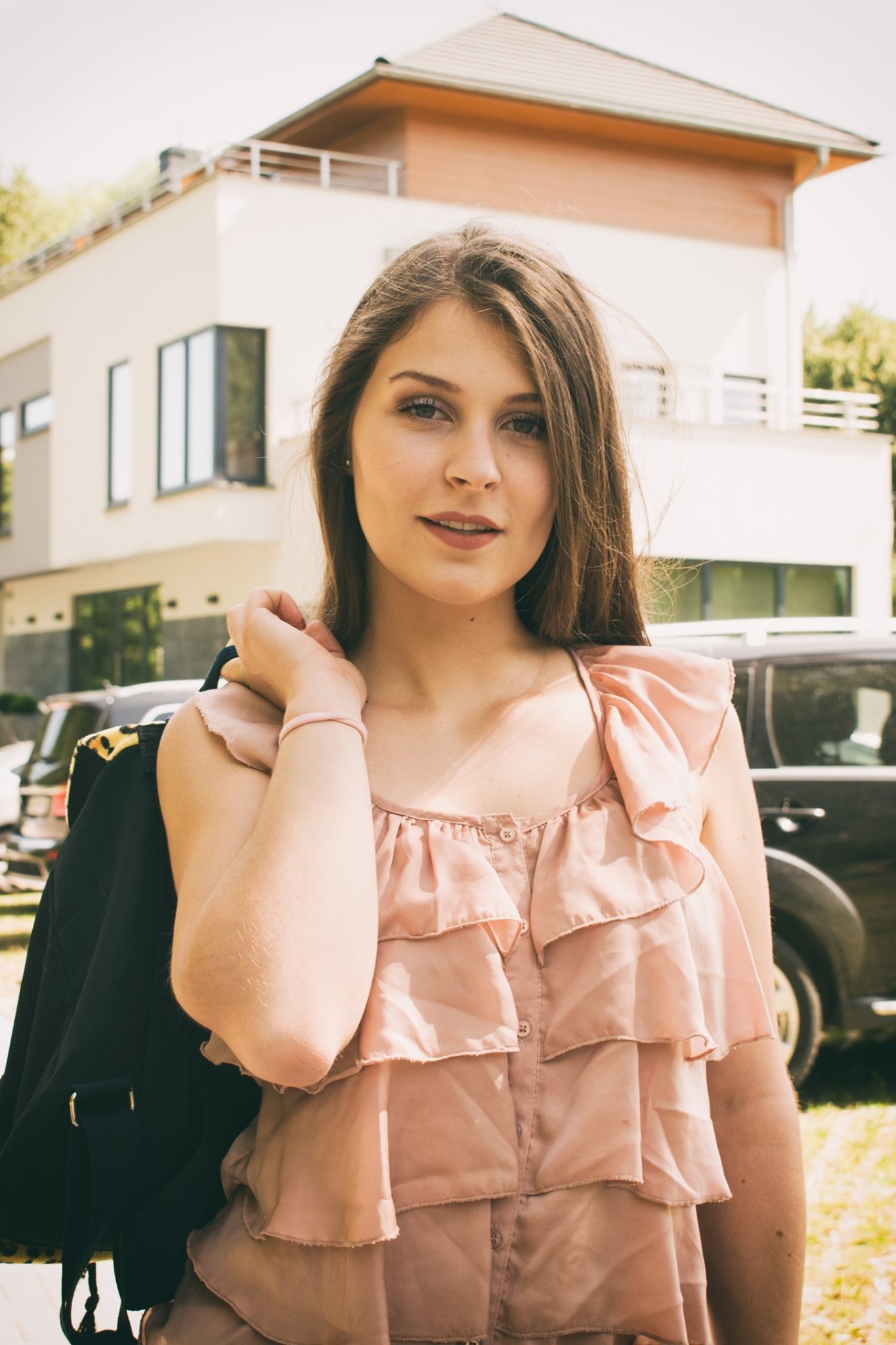 Portret młodej kobiety w bluzce z falbanami, na tle nowoczesnego domu i samochodu, z marynarką przewieszoną przez ramię.