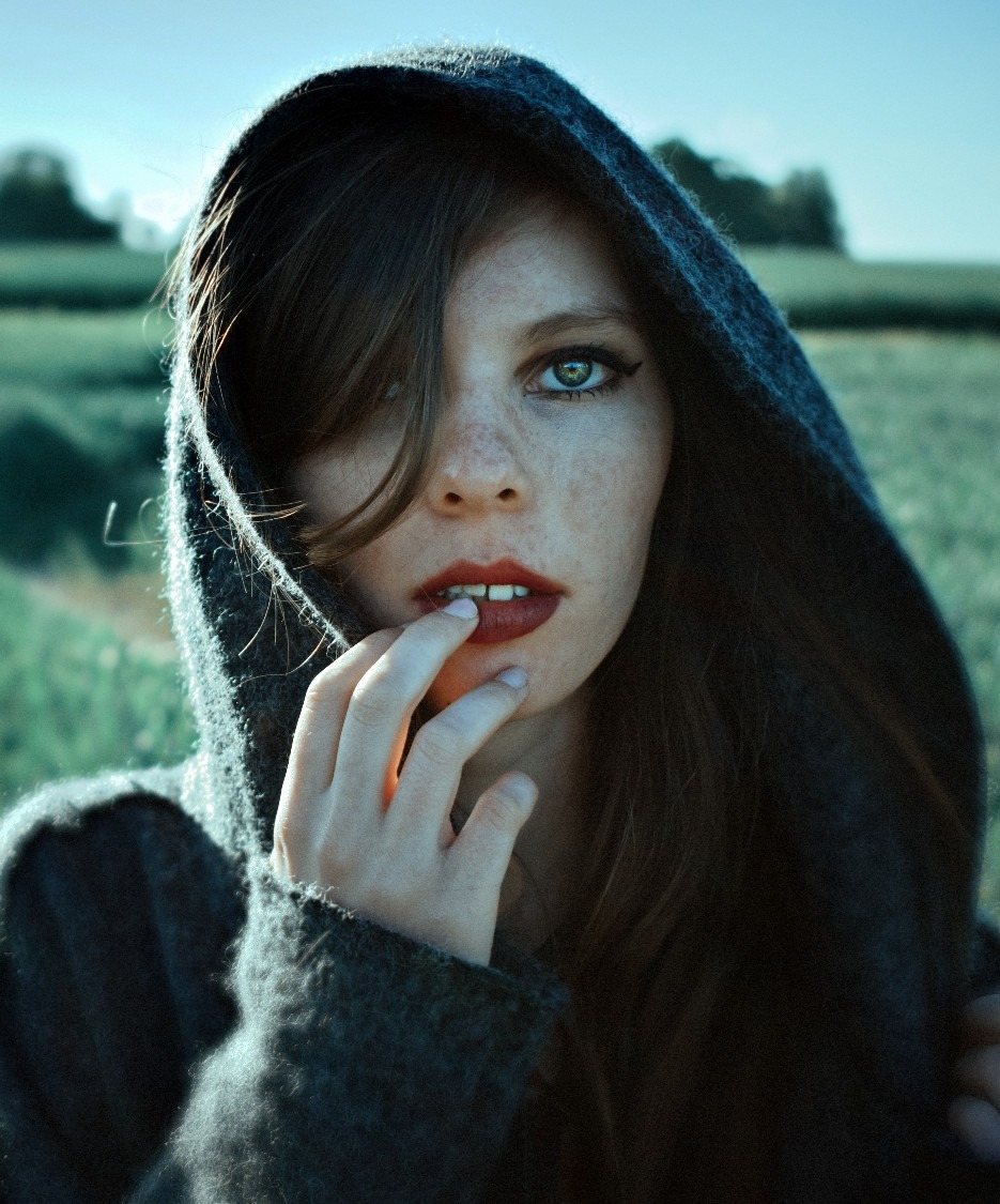 Portret młodej kobiety z piegami, w kapturze, z palcem przy ustach, na tle zielonego pola i błękitnego nieba.