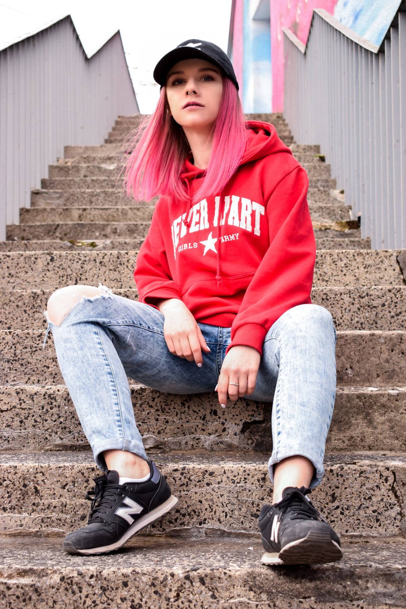 Młoda kobieta z różowymi włosami w czerwonej bluzie z kapturem i podartych jeansach siedzi na betonowych schodach w Gdyni, pozując do zdjęcia.