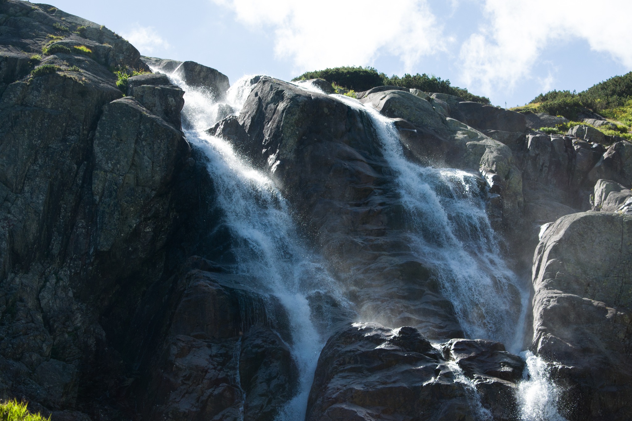 Kaskada wodna spływająca po skalistej górze, z fragmentami zieleni na szczycie i błękitnym niebem w tle.