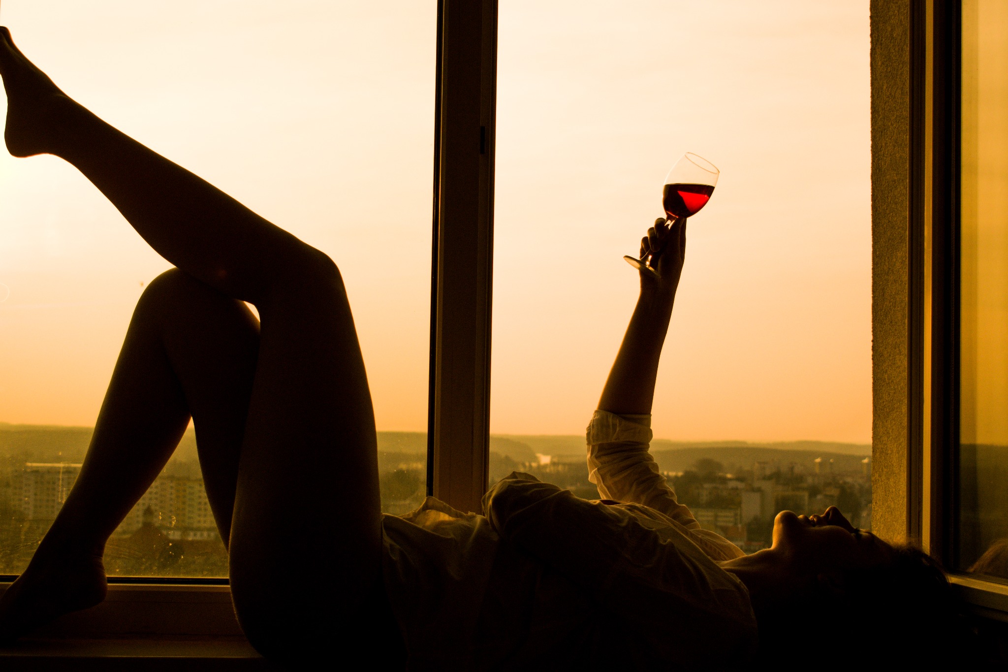 Sylwetka kobiety leżącej na parapecie z kieliszkiem czerwonego wina na tle zachodzącego słońca i widoku na miasto.