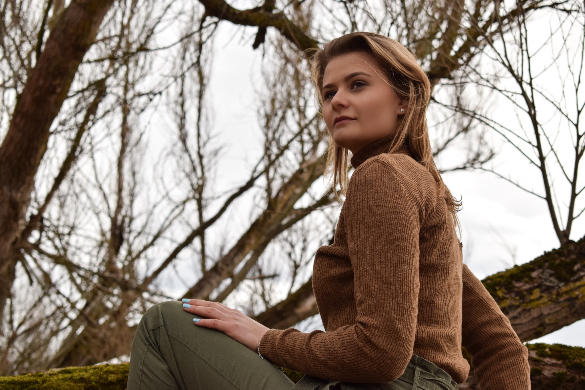 Młoda kobieta w brązowym swetrze i zielonych spodniach siedzi na gałęzi drzewa, spoglądając w dal; w tle widoczne nagie gałęzie drzew i pochmurne niebo.