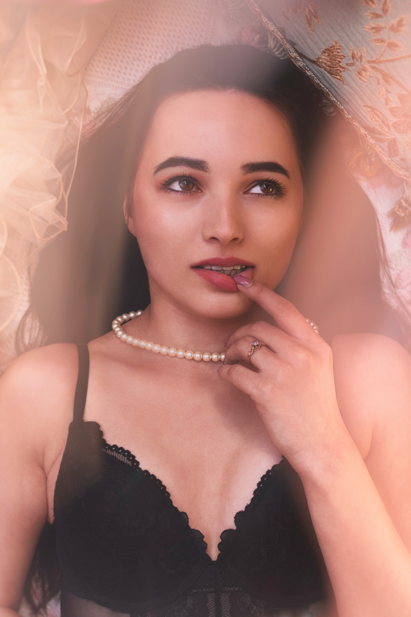 Kobieta w czarnej bieliźnie i perłach, z pierścionkiem na palcu, leży na wzorzystej tkaninie, spoglądając w górę z palcem przy ustach.