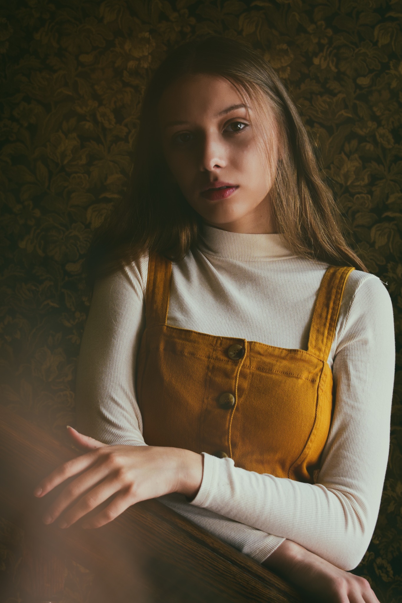 Portret młodej kobiety w musztardowej sukience na tle wzorzystej tapety, oświetlonej miękkim światłem.