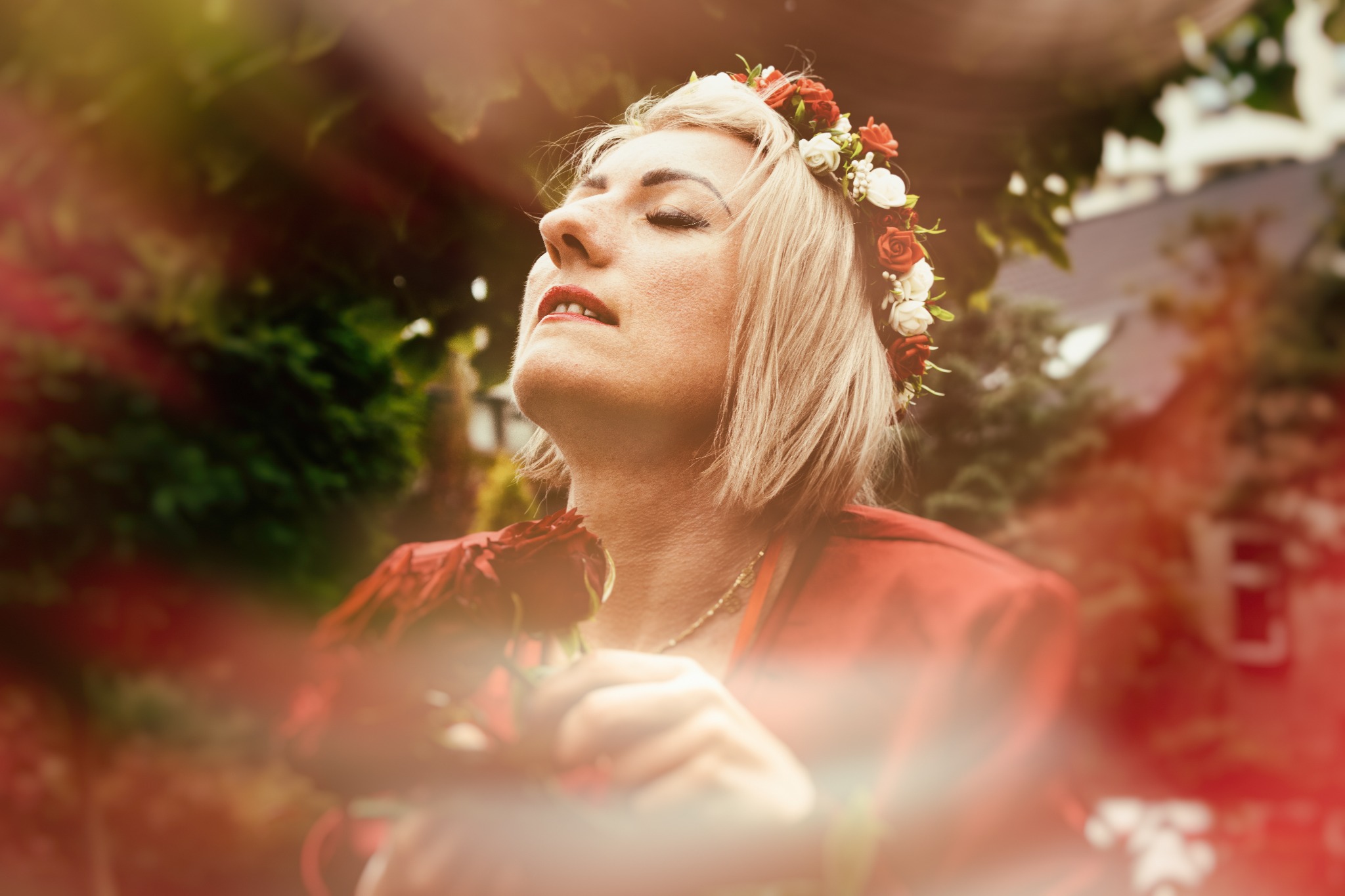 Kobieta z zamkniętymi oczami, w wianku z czerwonych i białych róż, trzymająca czerwoną różę, w kontraście z zielonym tłem.