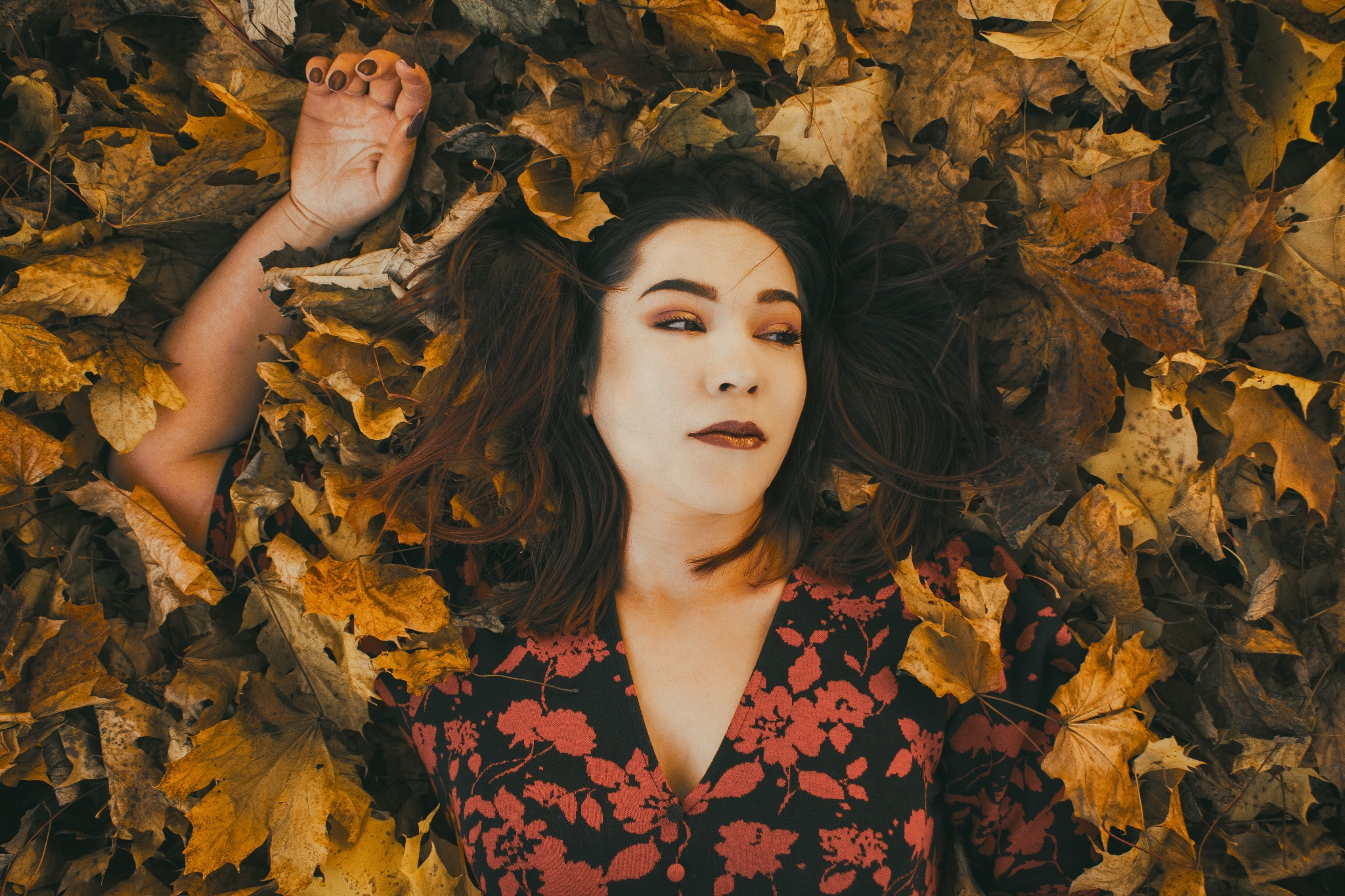 Kobieta leży w jesiennych liściach, ubrana w sukienkę w czerwone kwiaty na czarnym tle, z wyrazistym makijażem w odcieniach brązu i złota.