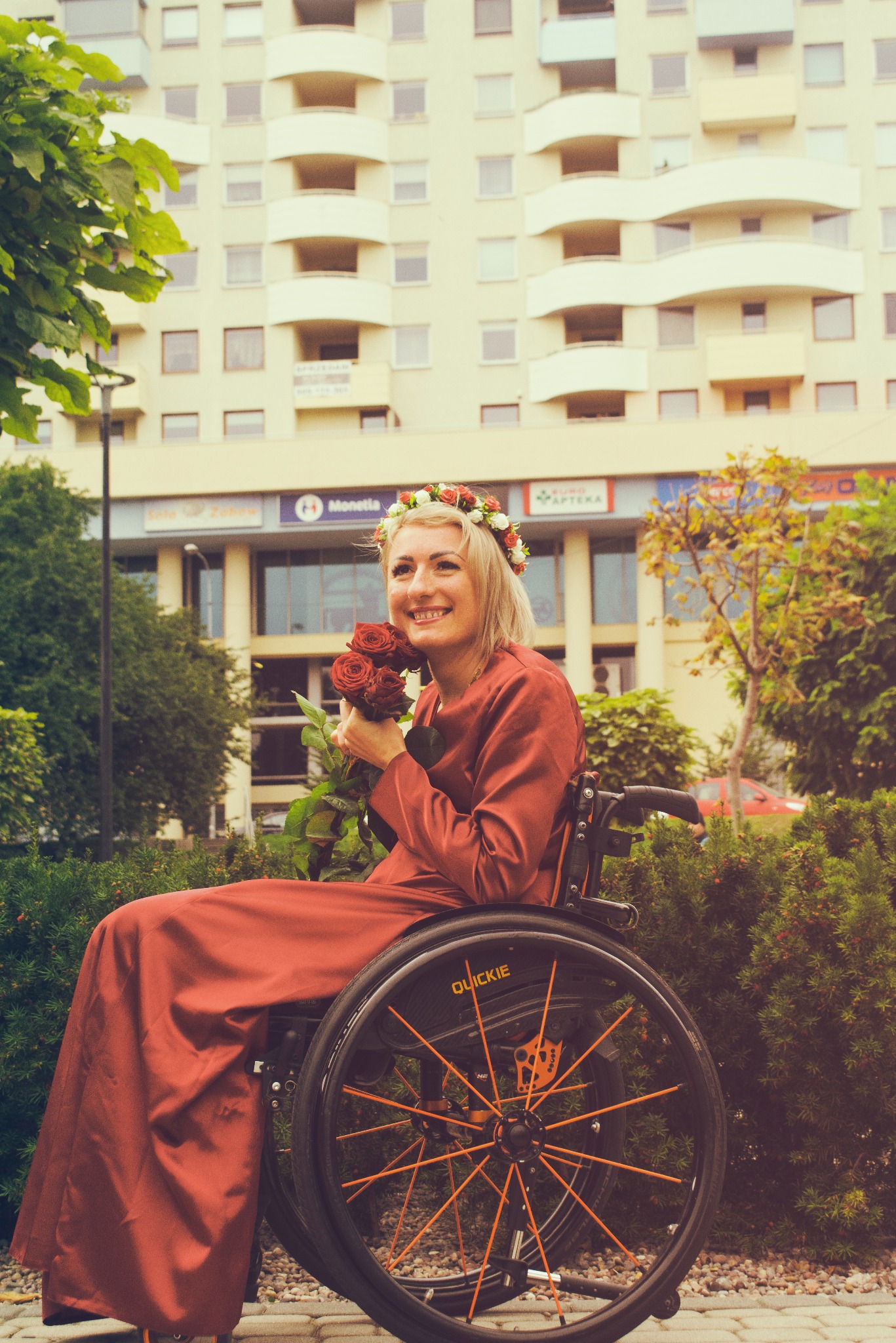 Uśmiechnięta blondynka w czerwonej sukni i wianku na głowie, siedząca na wózku inwalidzkim, trzyma bukiet czerwonych róż na tle budynku z balkonami.