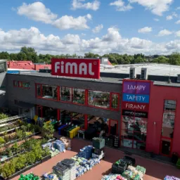 Widok z góry na budynek sklepu Fimal z ekspozycją roślin, ziemi ogrodowej w workach i kostki brukowej na zewnątrz, w tle zielone drzewa i błękitne niebo z białymi chmurami.