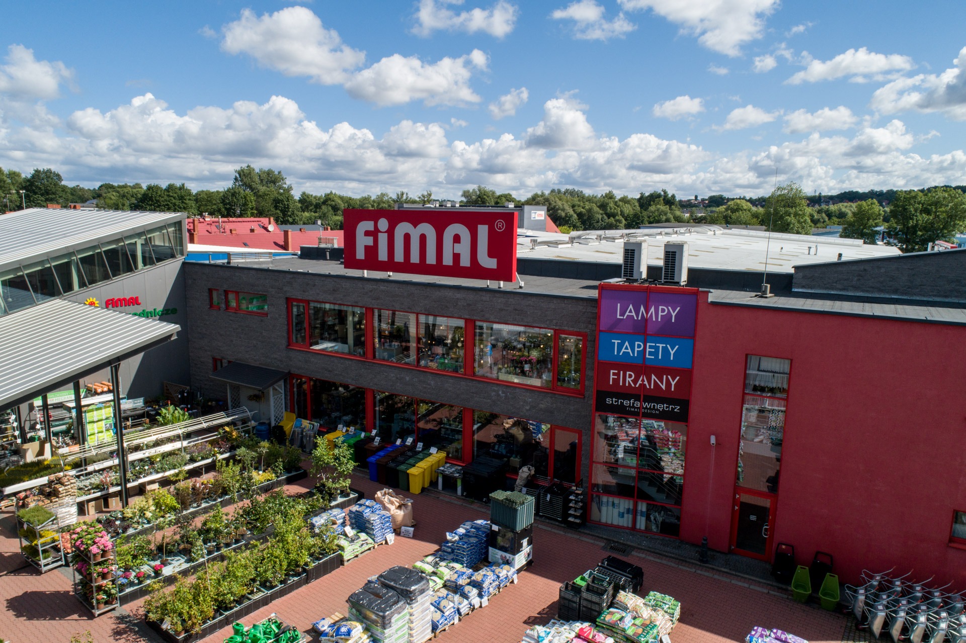 Widok z góry na budynek sklepu Fimal z ekspozycją roślin, ziemi ogrodowej w workach i kostki brukowej na zewnątrz, w tle zielone drzewa i błękitne niebo z białymi chmurami.