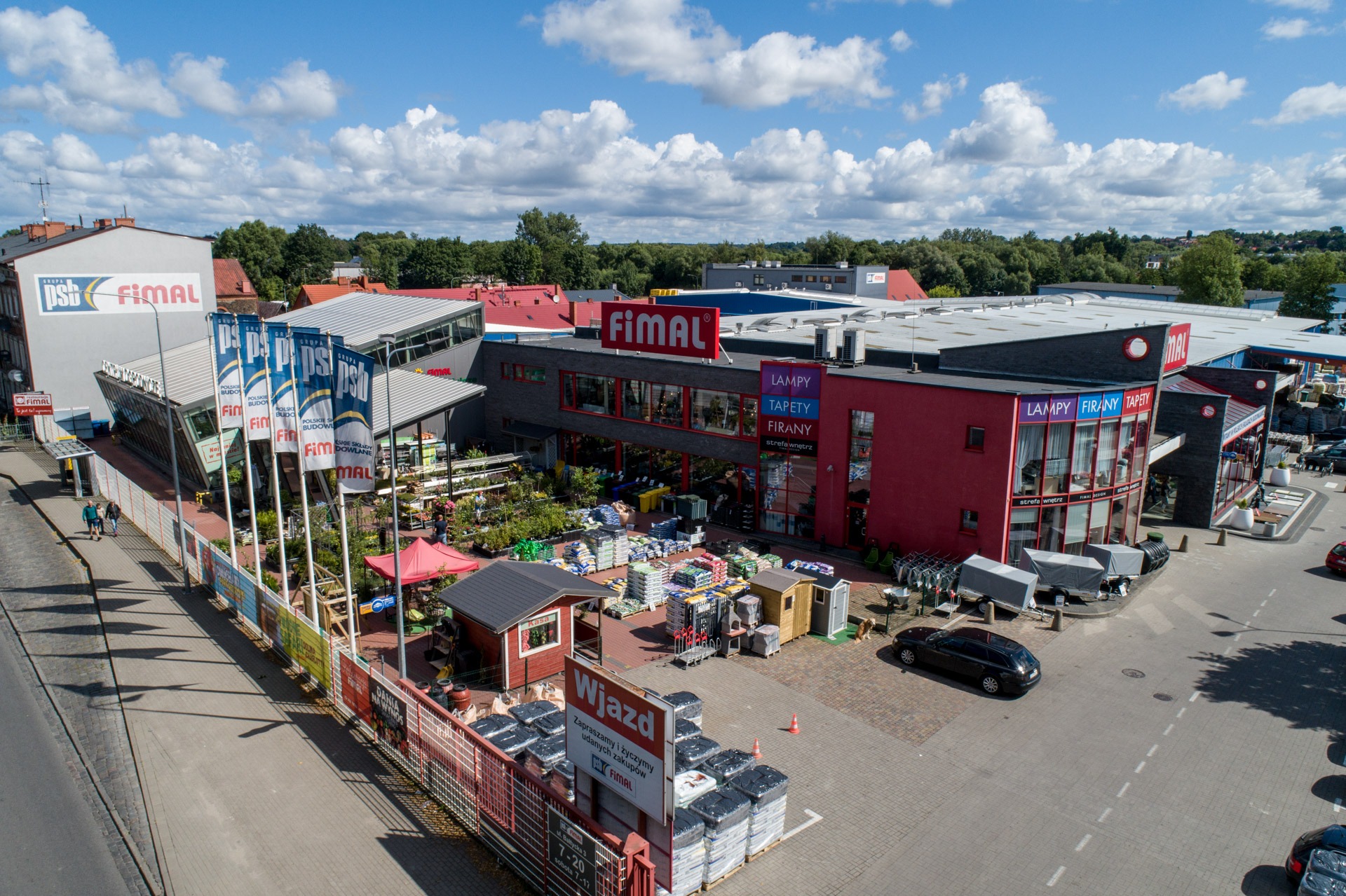 Szeroki widok z góry na teren składu budowlanego Fimal w słoneczny dzień, z widocznymi materiałami budowlanymi, roślinami ogrodowymi, budynkami z logo firmy i zaparkowanymi samochodami.