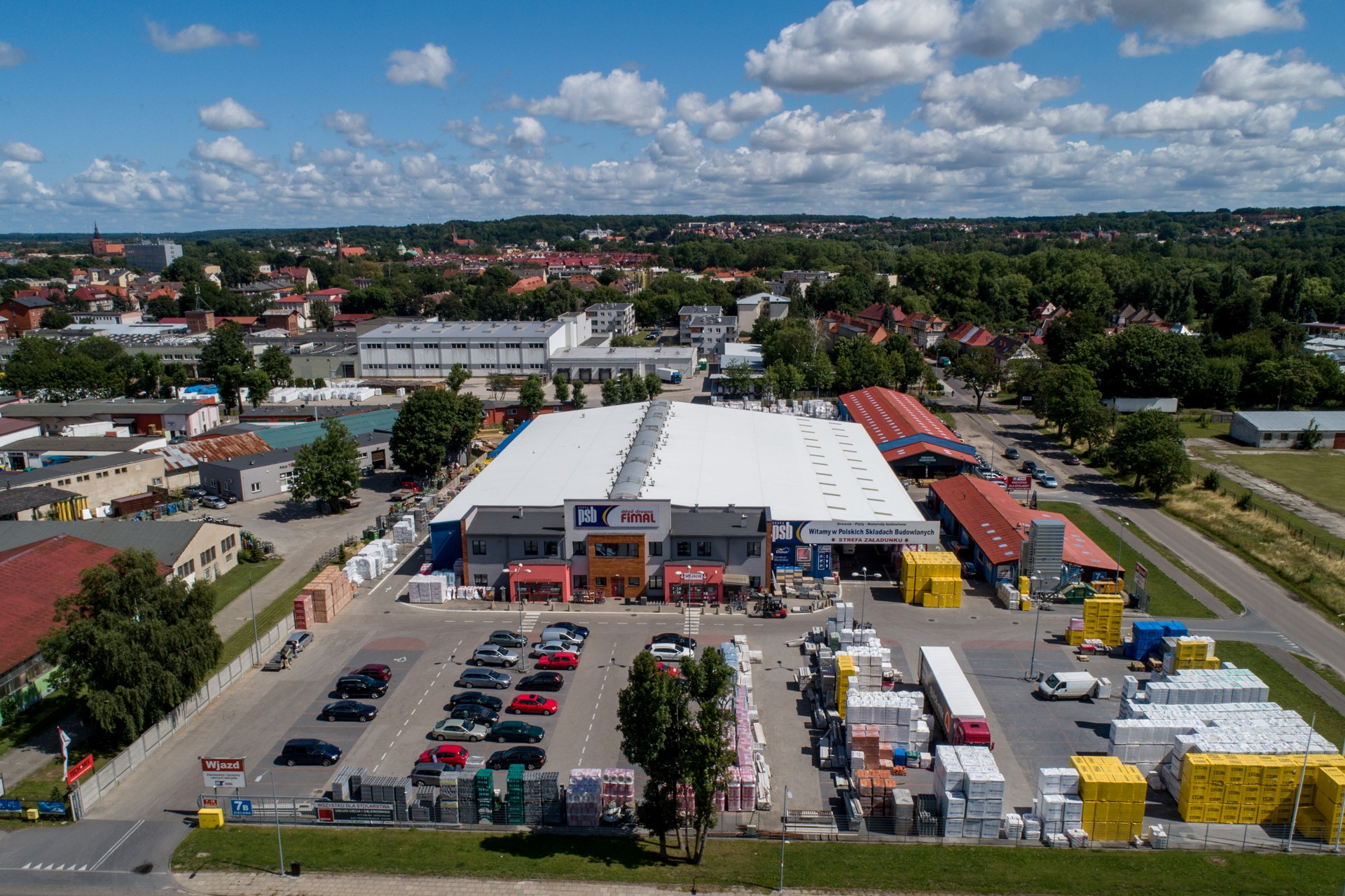 Szeroki widok z lotu ptaka na teren dużego składu budowlanego z zaparkowanymi samochodami i składowanymi materiałami budowlanymi, w tle zabudowa miejska i zalesione wzgórza pod błękitnym niebem...