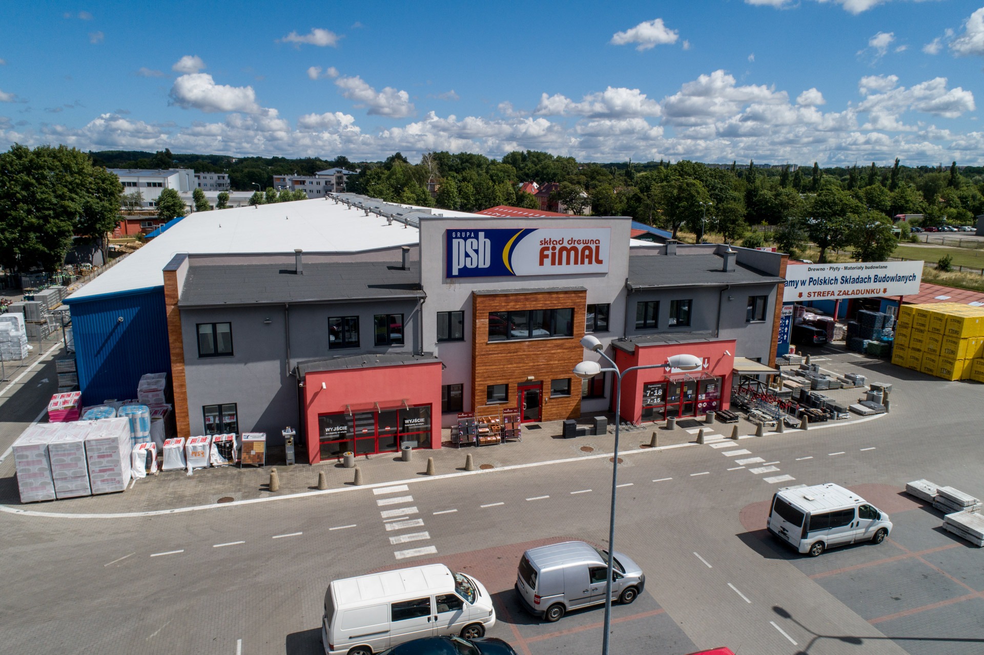 Szeroki kadr z lotu ptaka na budynek składu materiałów budowlanych Grupa PSB FIMAL z zaparkowanymi samochodami dostawczymi i ułożonymi paletami z towarem na zewnątrz.