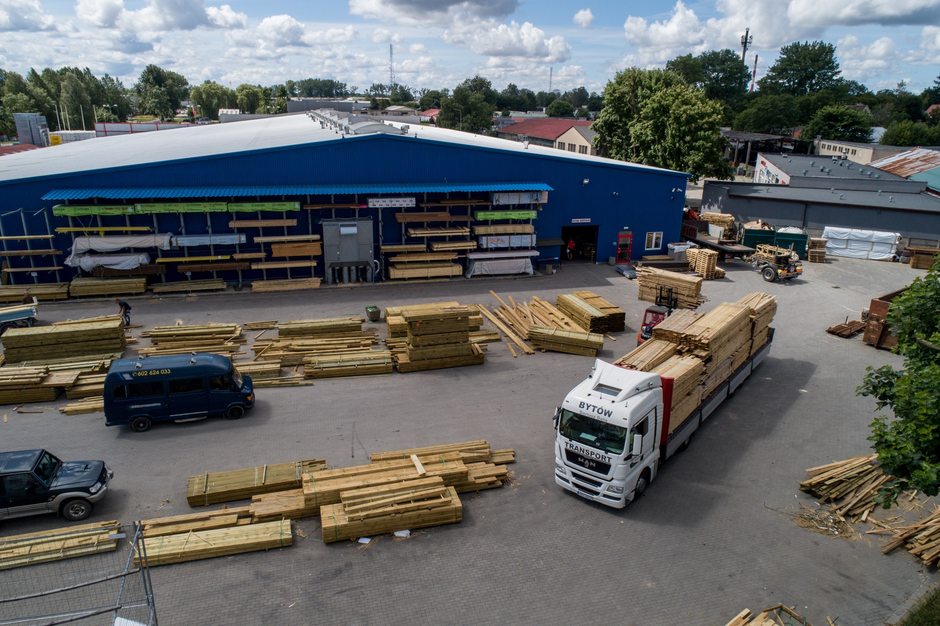 Widok z góry na teren składu z zapasem drewna budowlanego, ciężarówką transportową i pracującym człowiekiem przy rozładunku.