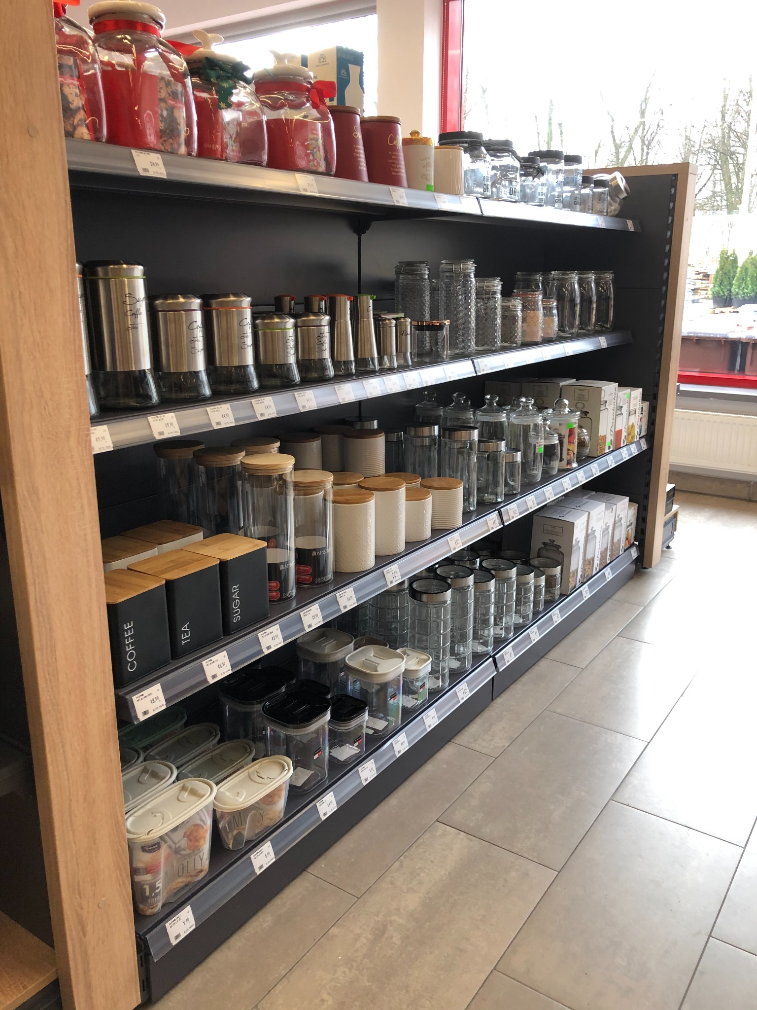 Półki sklepowe wypełnione różnorodnymi pojemnikami do przechowywania żywności, w tym szklanymi słoikami z drewnianymi pokrywkami, metalowymi puszkami na kawę i herbatę oraz plastikowymi pojemnikami...