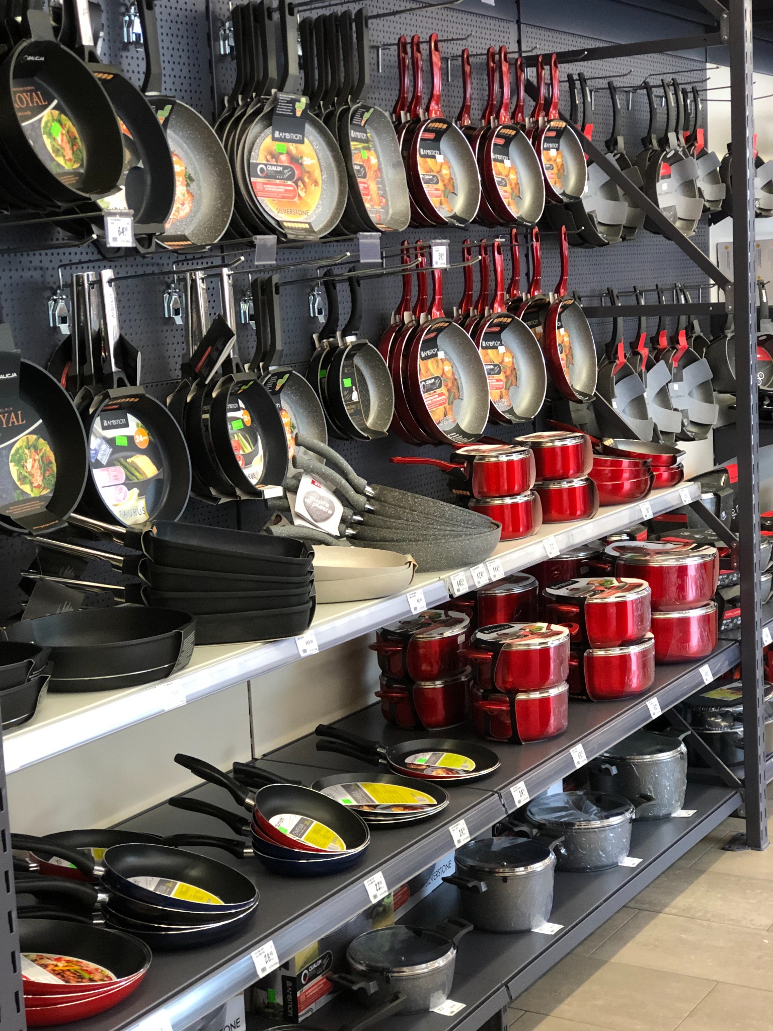 Ekspozycja różnorodnych garnków i patelni marek Ambition i Royal na półkach sklepowych, widoczne różne kolory i rozmiary, od patelni z powłoką Silverstone po czerwone komplety garnków.