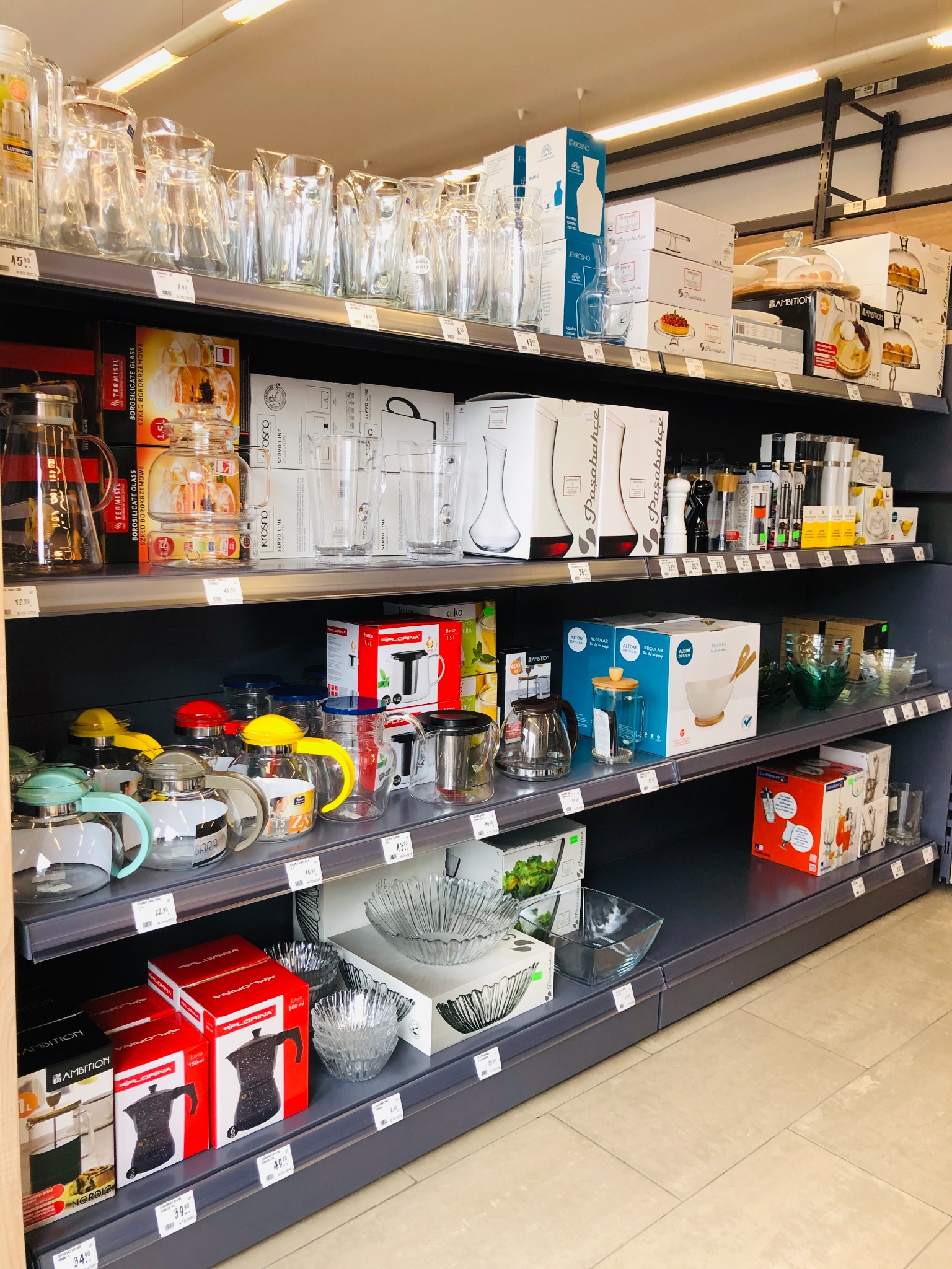 Półki sklepowe z różnorodnym asortymentem szklanych i metalowych naczyń kuchennych, w tym dzbanki, szklanki, miski i kawiarki, prezentowane w opakowaniach i bez, z widocznymi cenami.