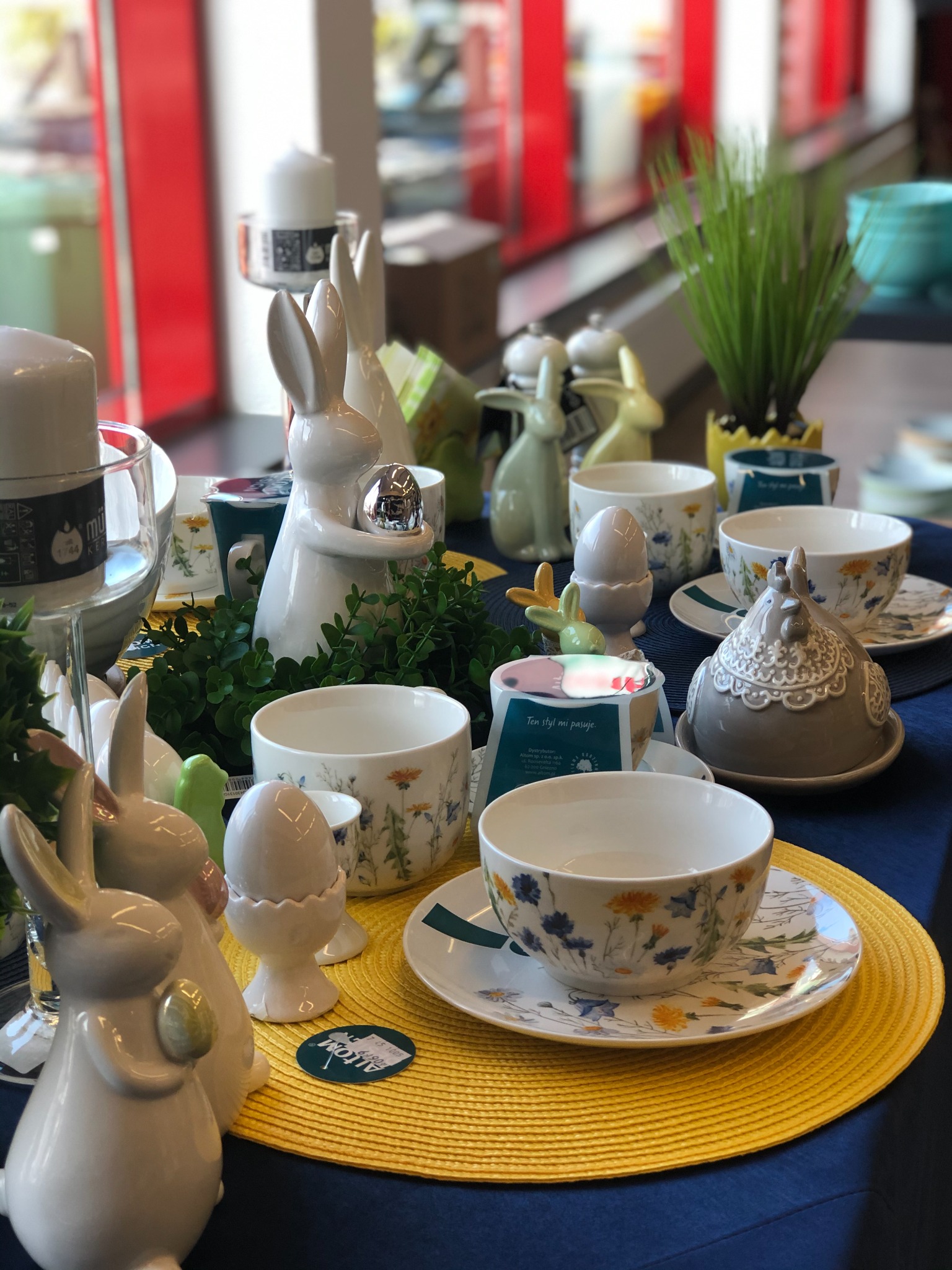 Aranżacja stołu z dekoracjami wielkanocnymi: ceramiczne zajączki, miseczki z motywem kwiatowym, podstawki na jajka i zielone rośliny na żółtej podkładce.