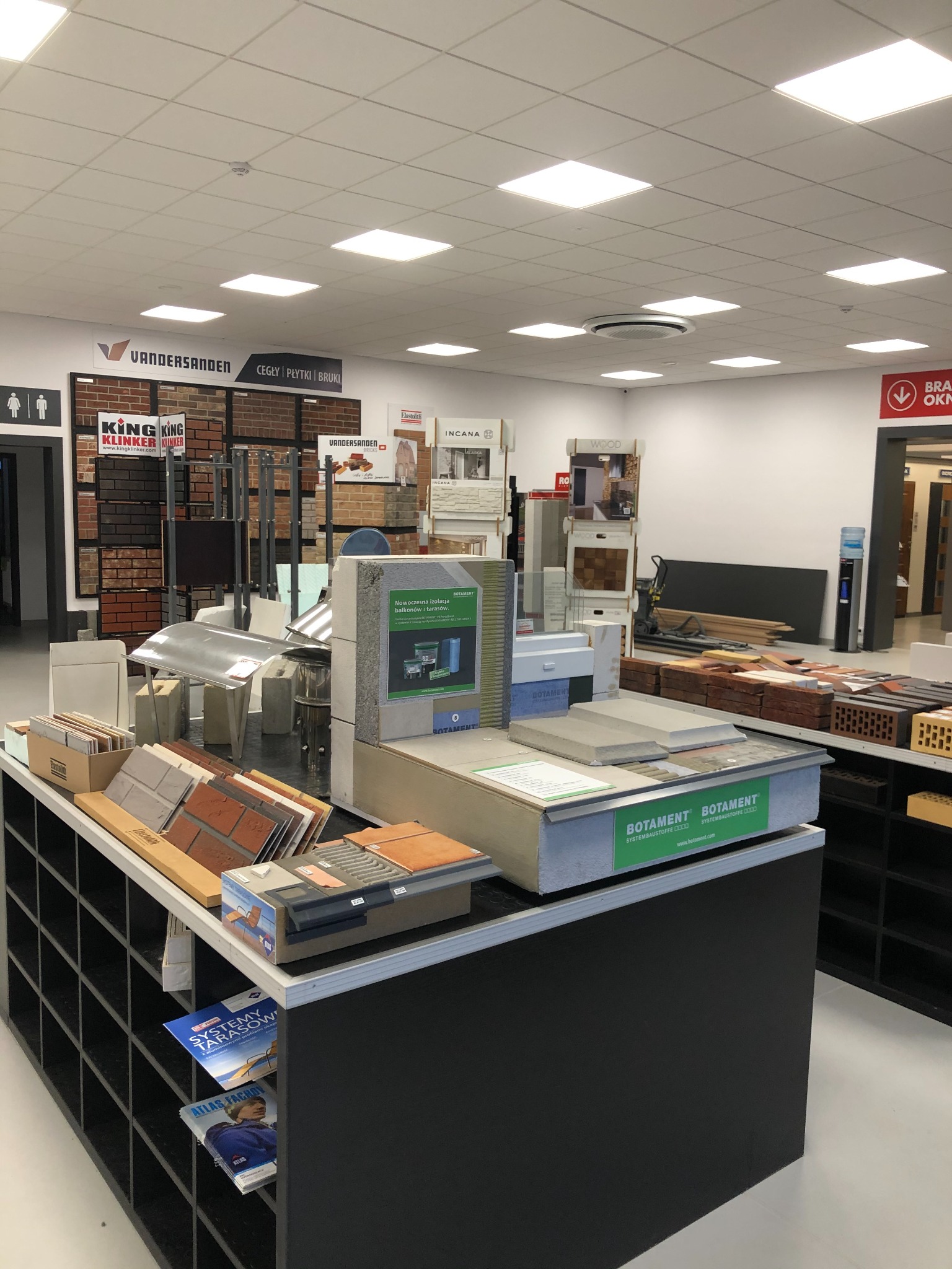 Ekspozycja materiałów budowlanych wewnątrz sklepu: cegły klinkierowe, płytki elewacyjne, systemy izolacji balkonów i tarasów marki Botament, katalogi reklamowe, oświetlenie sufitowe.