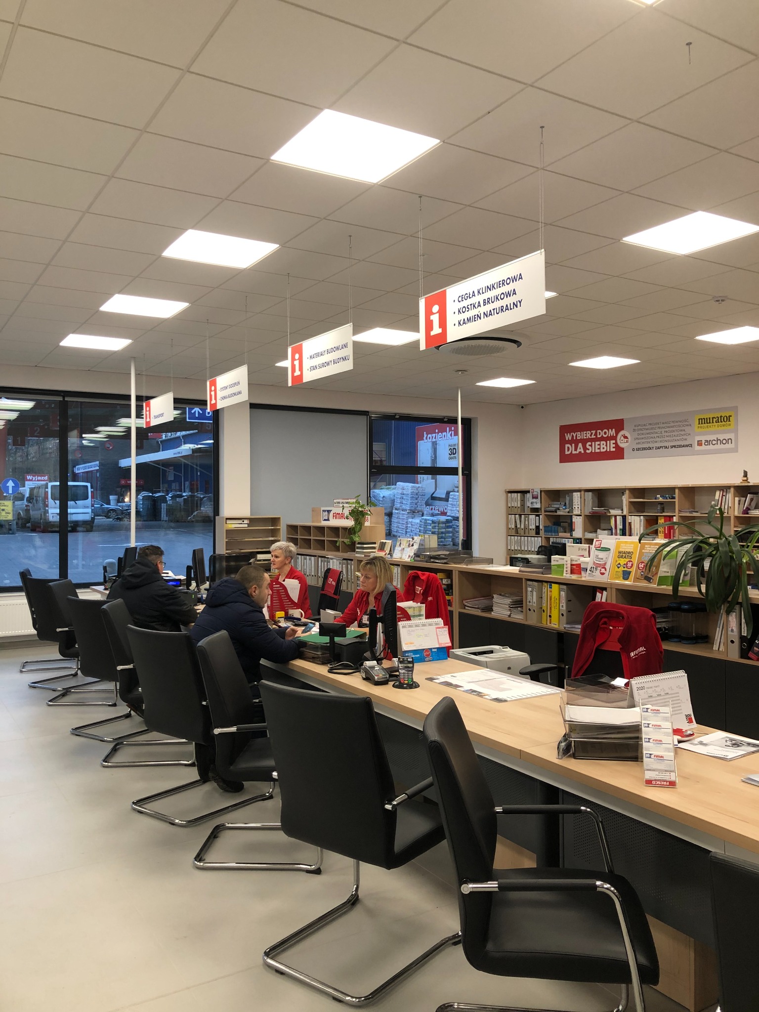 Wnętrze punktu obsługi klienta w sklepie budowlanym z pracownikami przy biurkach, wiszące tablice informacyjne o materiałach budowlanych i kostce brukowej, widok na zewnątrz z zapasami materiałów.