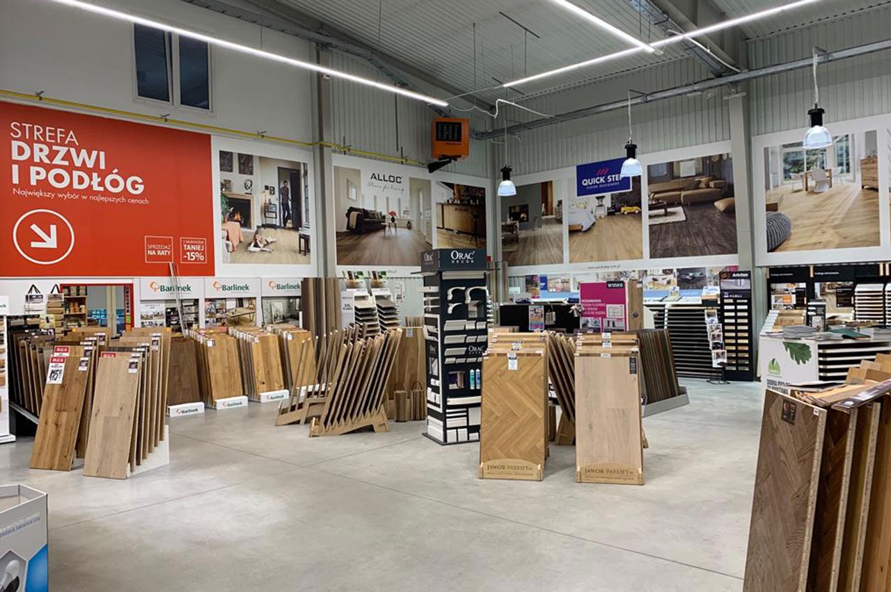 Wnętrze sklepu z ekspozycją paneli podłogowych różnych marek i wzorów, plakaty reklamowe na ścianach, oświetlenie sufitowe.