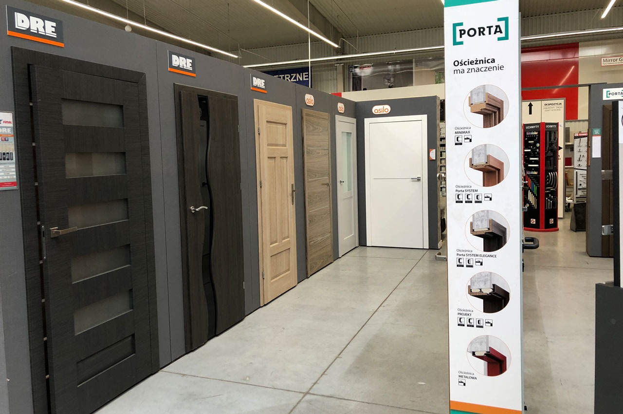 Ekspozycja różnorodnych drzwi wewnętrznych marek DRE, Asilo i Porta w sklepie, prezentująca różne modele, kolory i wykończenia, w tym drzwi z przeszkleniami i panel reklamowy Porta.
