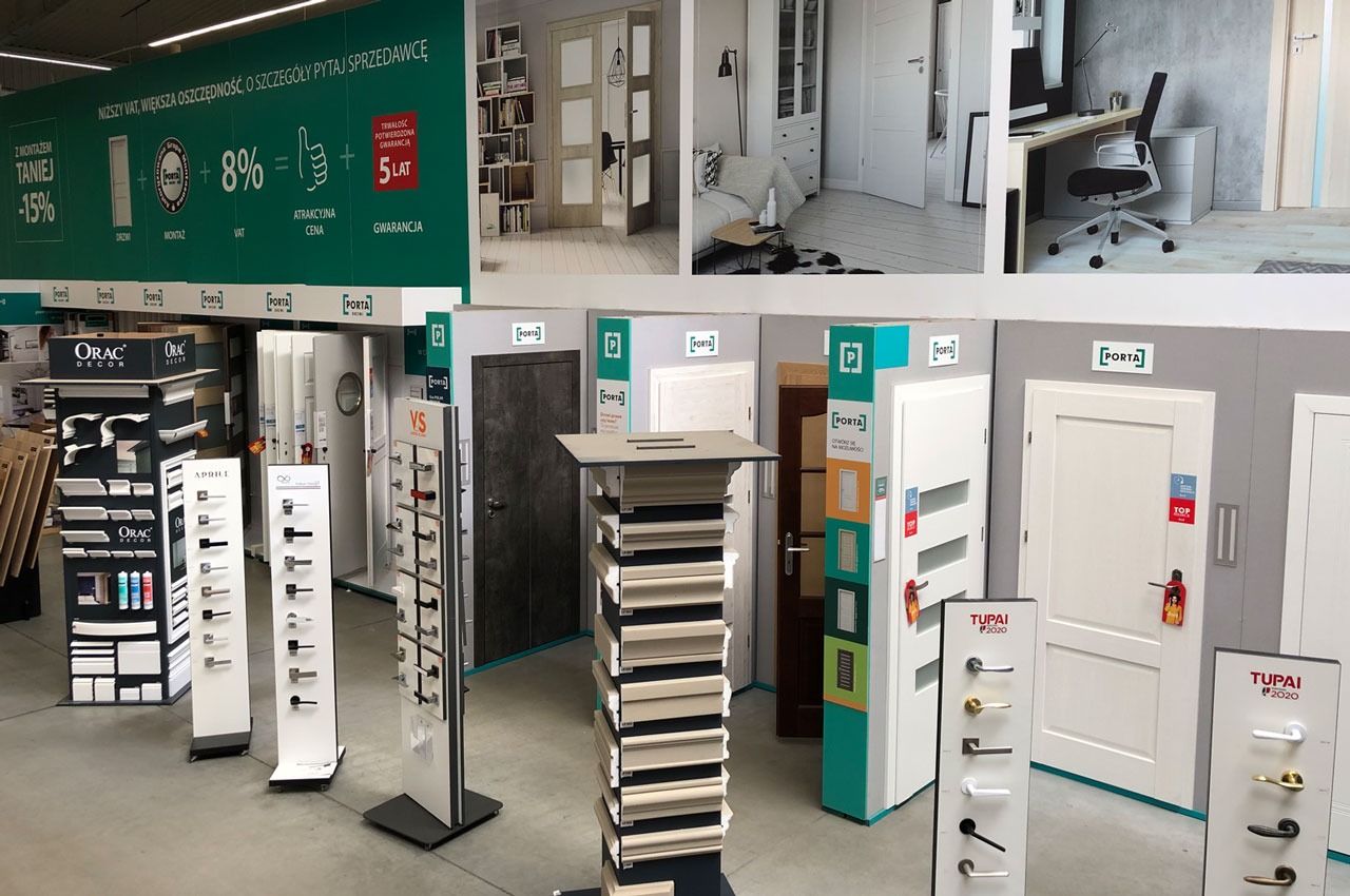 Ekspozycja różnych modeli drzwi wewnętrznych i klamek w sklepie, widoczne także reklamy promocyjne.