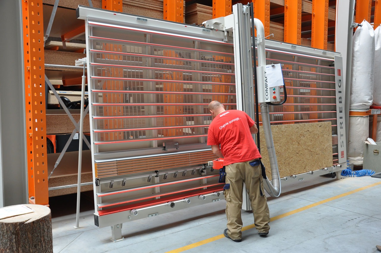 Pracownik w czerwonej koszulce obsługuje pionową piłę panelową STRIEBIG Compact, tnąc płytę OSB w magazynie z pomarańczowymi regałami wypełnionymi płytami.