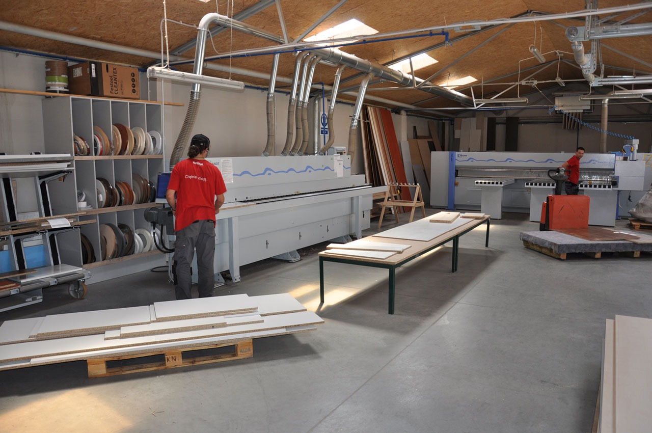 Produkcja elementów meblowych w hali: pracownicy obsługują maszyny do obróbki drewna, widoczne płyty meblowe na palecie i stole, system odciągu pyłu pod sufitem.