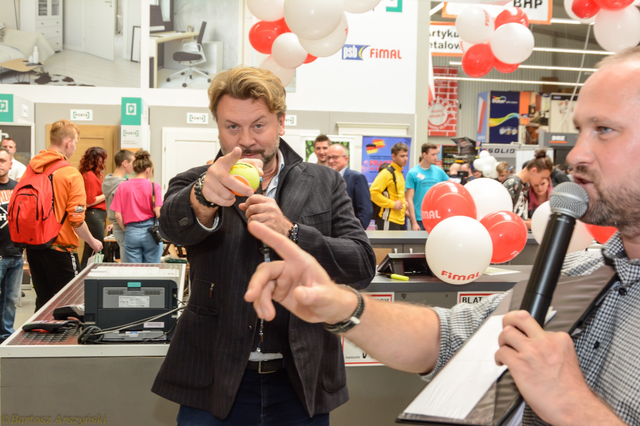 Mężczyzna w garniturze wskazuje palcem trzymając piłeczkę tenisową, obok mężczyzna z mikrofonem na tle sklepu budowlanego z balonami i klientami.
