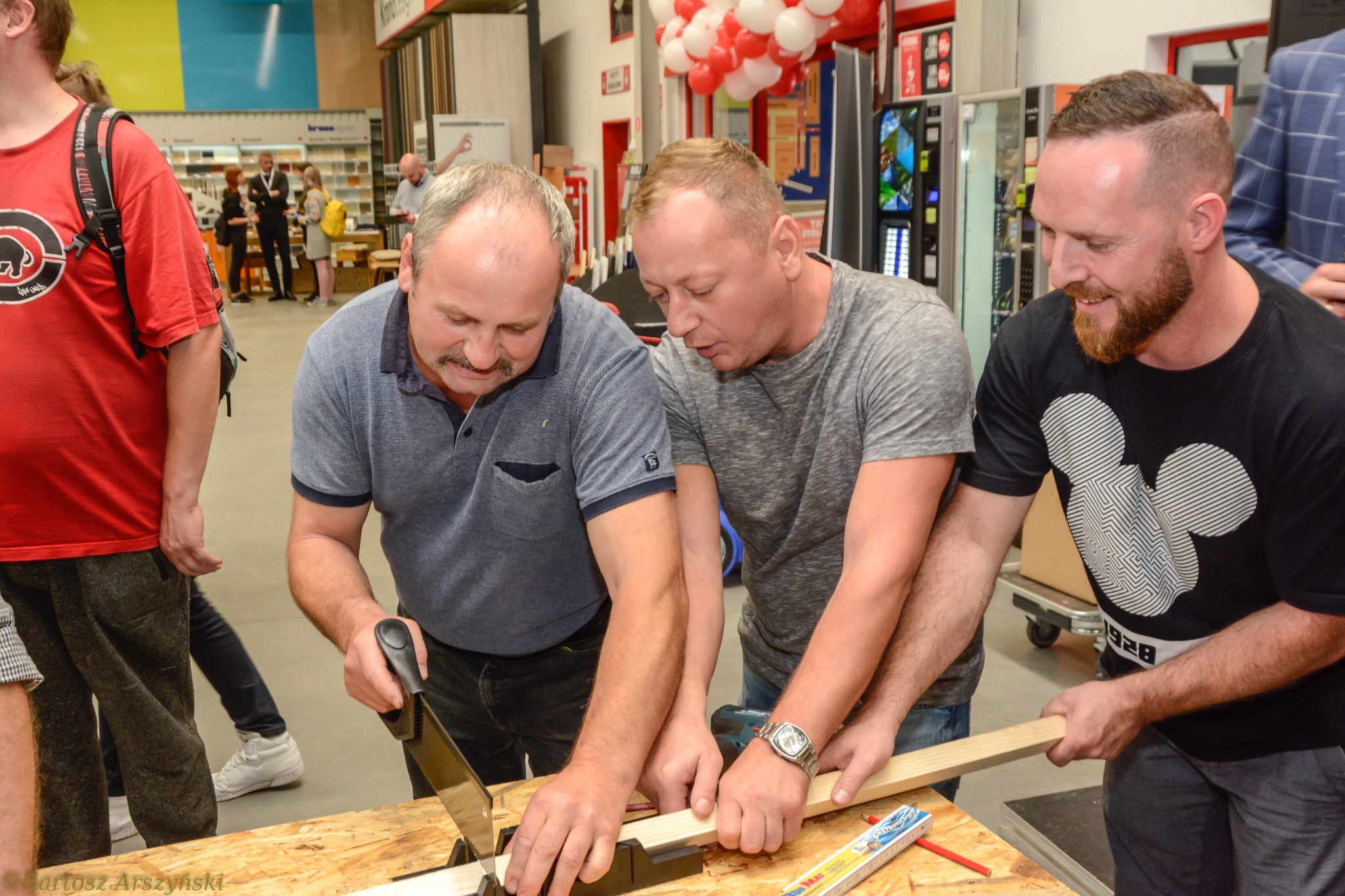 Trzech mężczyzn przycinających drewnianą listwę za pomocą piły ręcznej w skrzynce uciosowej w sklepie budowlanym. Jeden z nich ma na sobie koszulkę z motywem Myszki Miki.