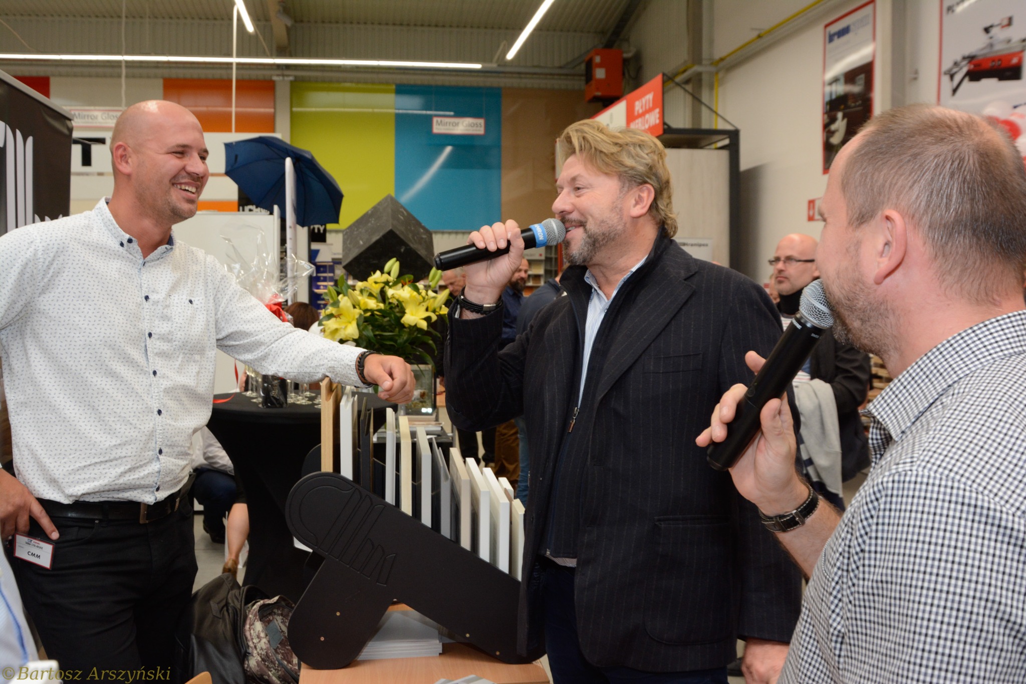 Prezenter z mikrofonem przemawia podczas prezentacji próbek materiałów budowlanych na targach, w tle uśmiechnięty mężczyzna i inne osoby.