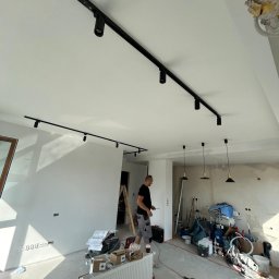 Cirqo Instalacje Elektryczne Bartłomiej Musiał - Montaż szynoprzewodów z reflektorami w nowym mieszkaniu, widoczny elektryk z narzędziami, surowy stan wykończenia wnętrza, okablowanie zwisające z sufitu w tle.